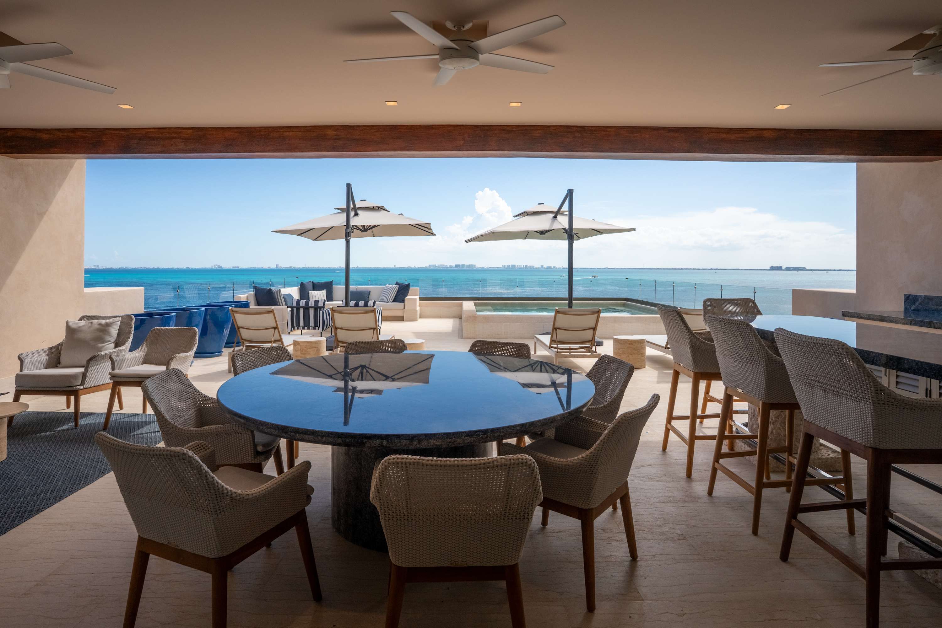 A covered outdoor patio with a dining table, chairs, and a bar overlooking a turquoise ocean.