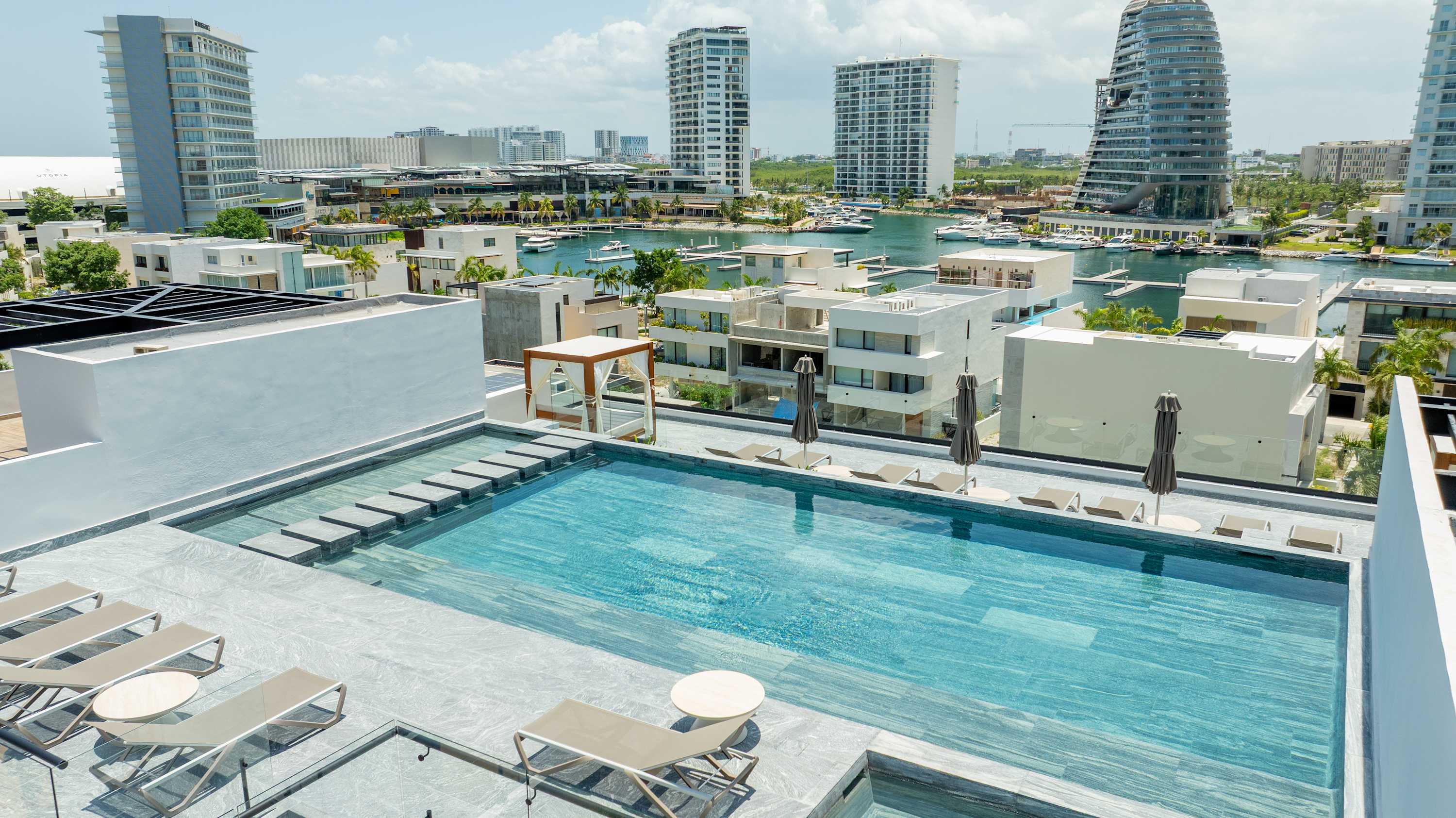 Rooftop pool with lounge chairs and umbrellas overlooking a harbor with boats and modern buildings.