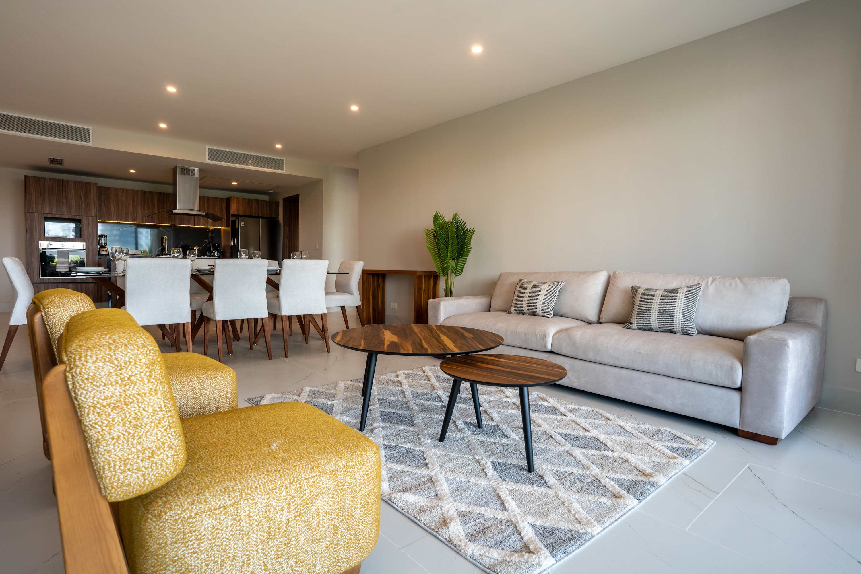 Modern living room with a light gray sofa, yellow chairs, dining table, and kitchen.