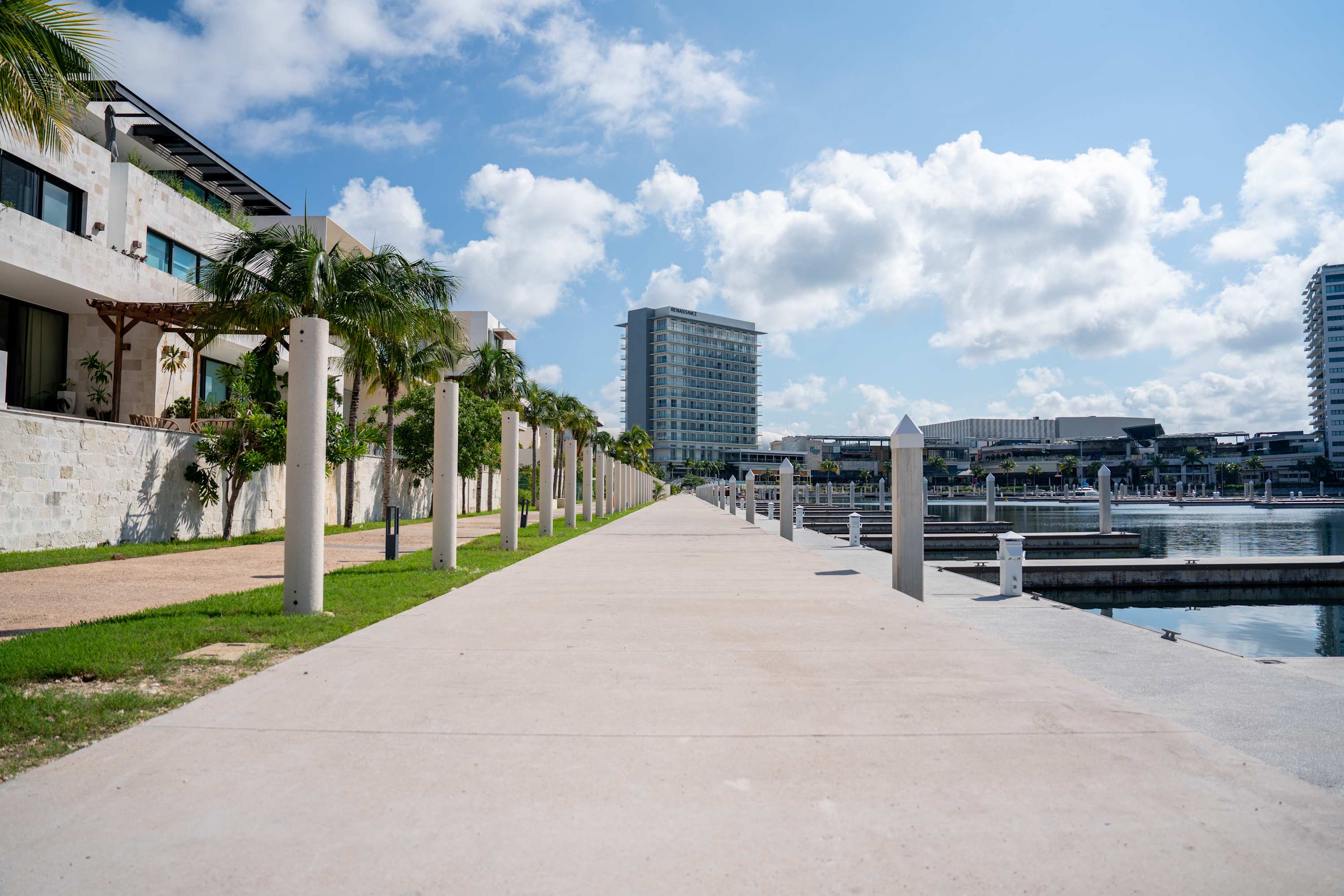A long concrete walkway stretches towards buildings and a marina under a cloudy sky.