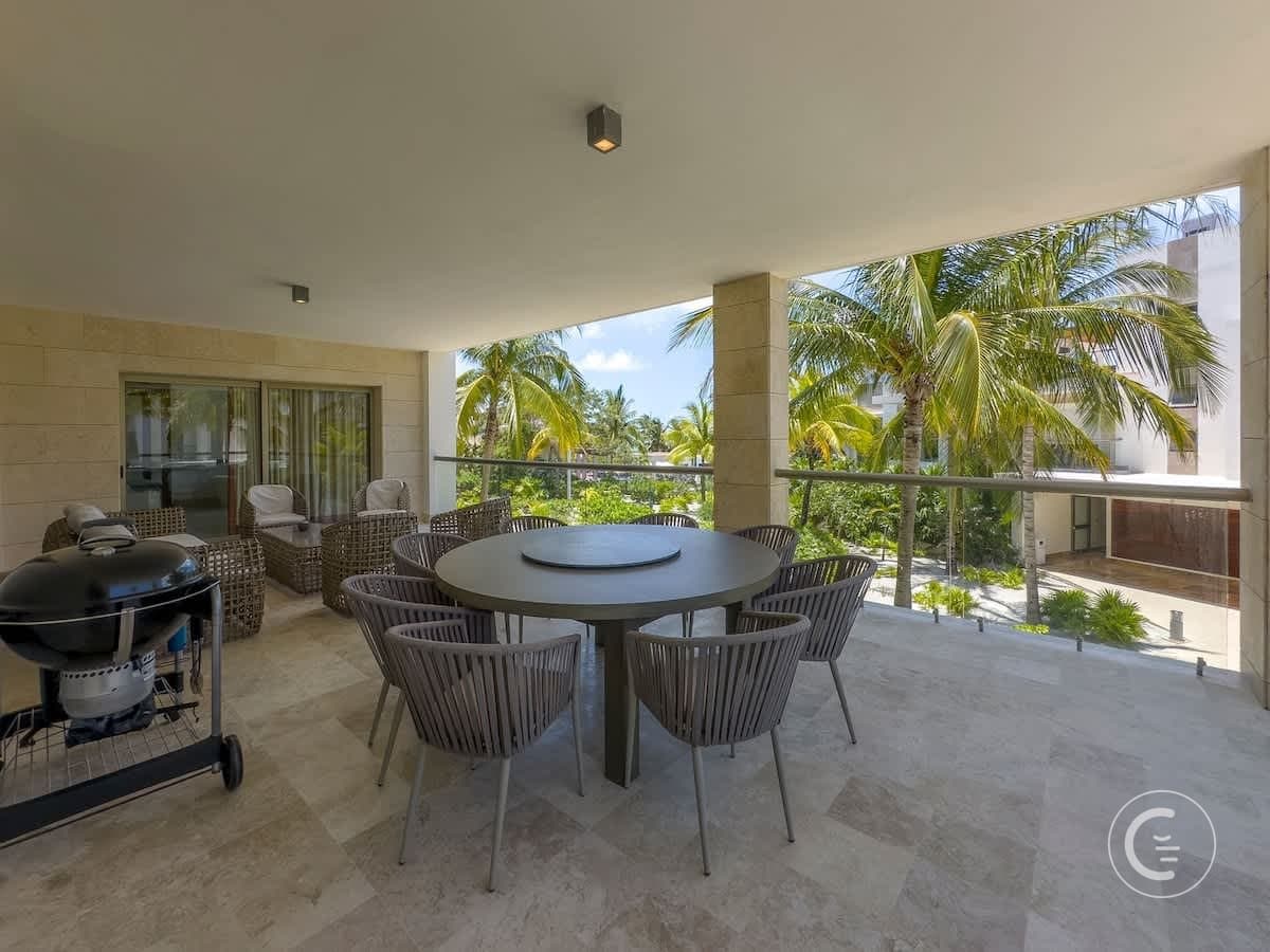 A spacious balcony with a round table and chairs, a grill, and palm trees in the background.