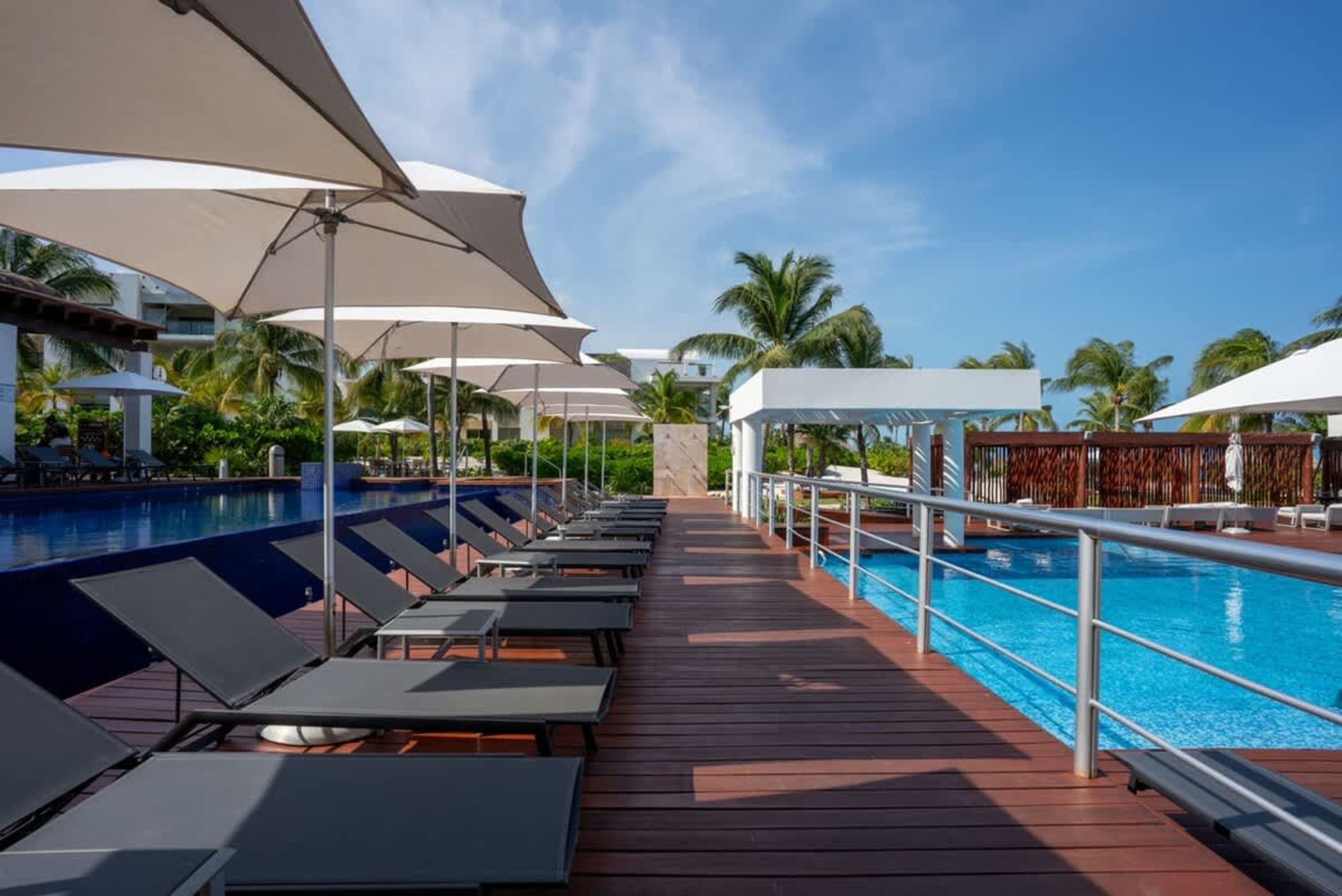 A wooden deck with lounge chairs and umbrellas surrounds a bright blue swimming pool.