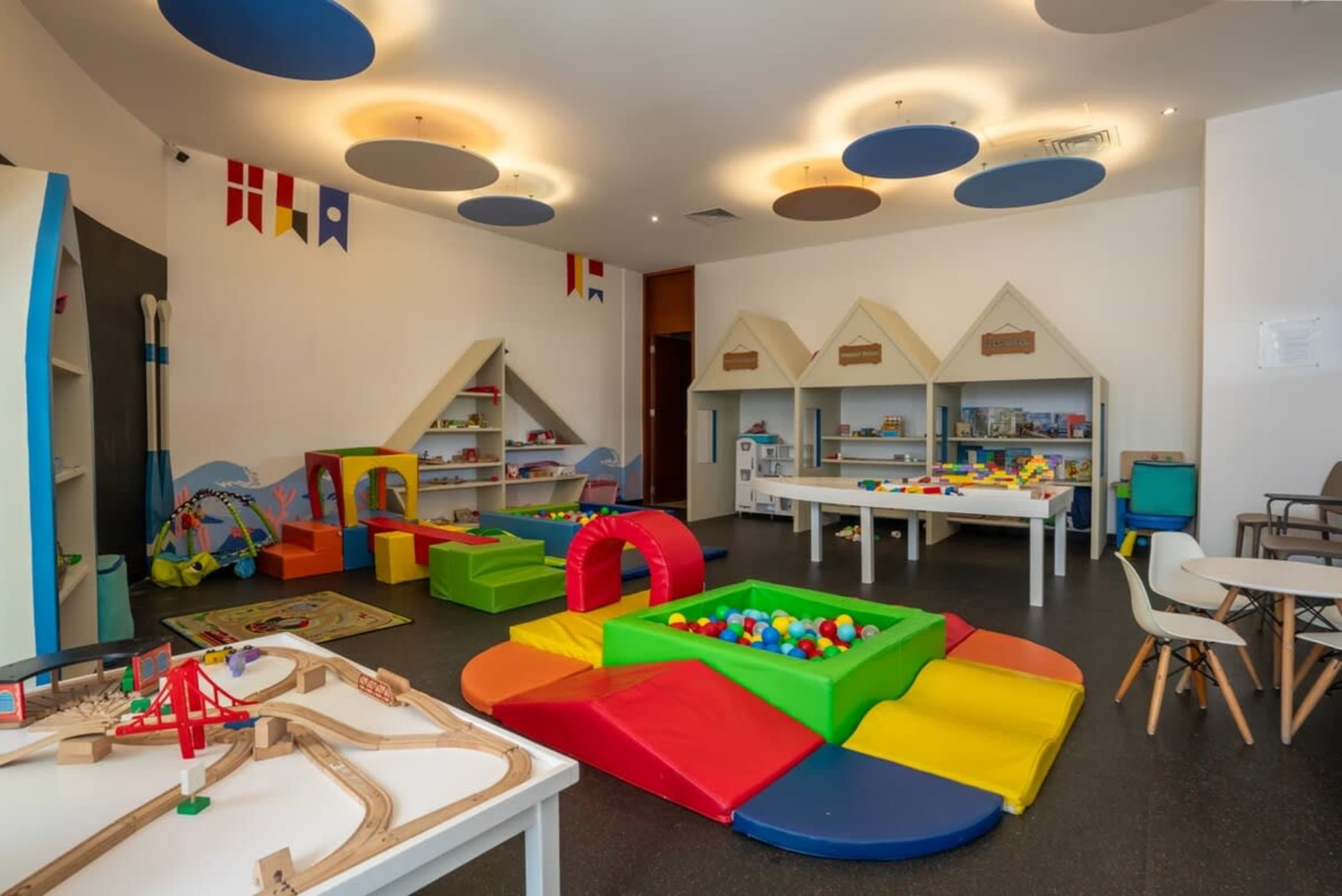 A colorful playroom with soft play structures, ball pit, toy shelves, and a wooden train set.