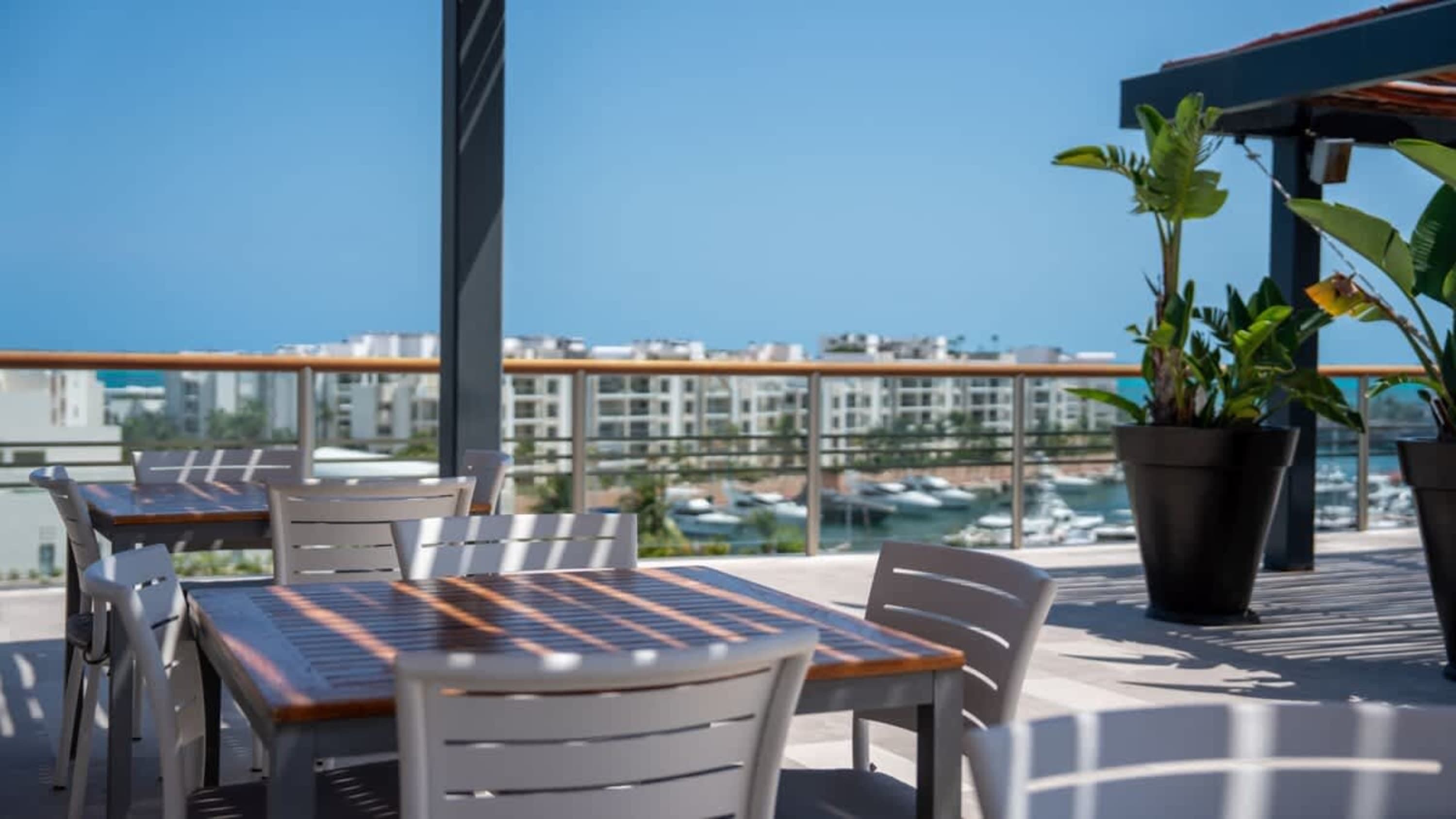 An outdoor dining area with tables and chairs overlooks a harbor with boats and buildings.