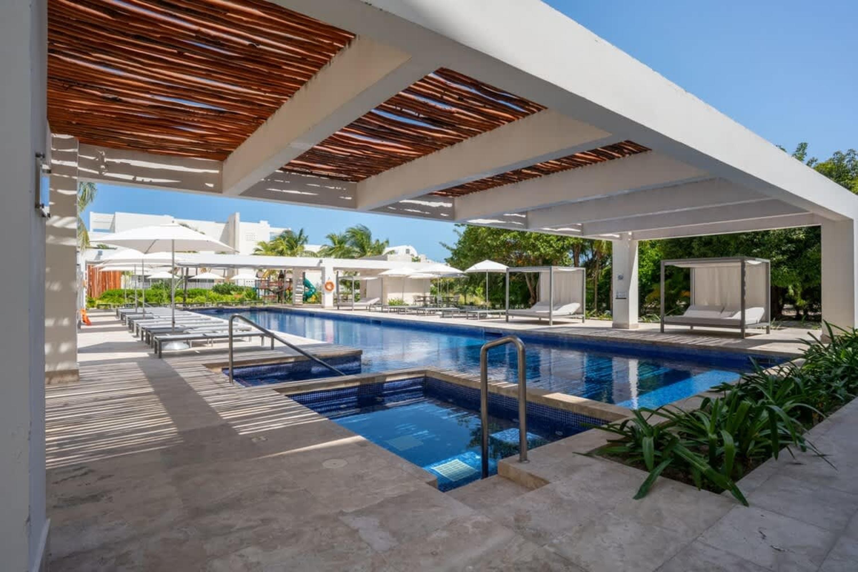 A resort pool area with lounge chairs and cabanas under a wooden beamed canopy.