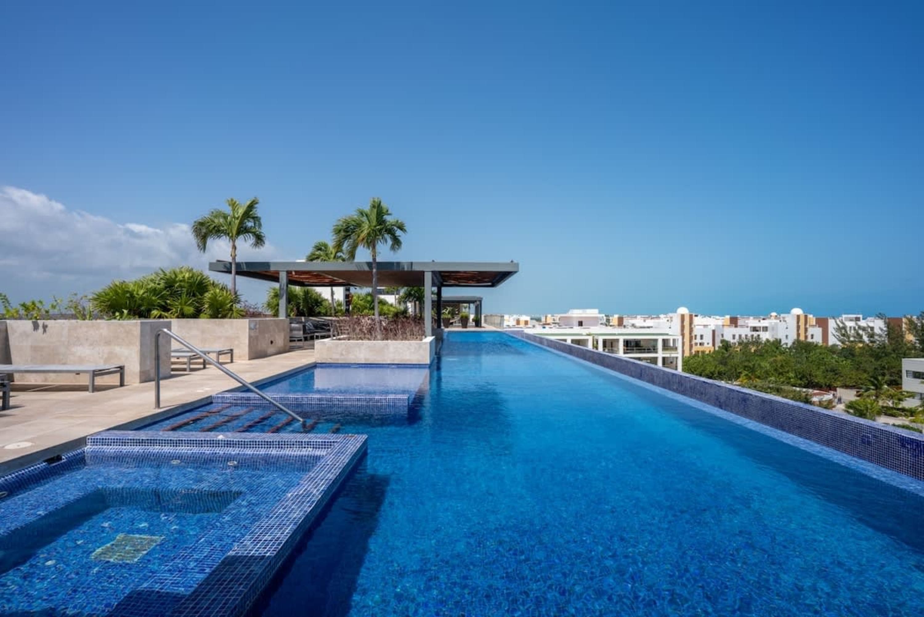Rooftop infinity pool with lounge chairs, palm trees, and a city view.