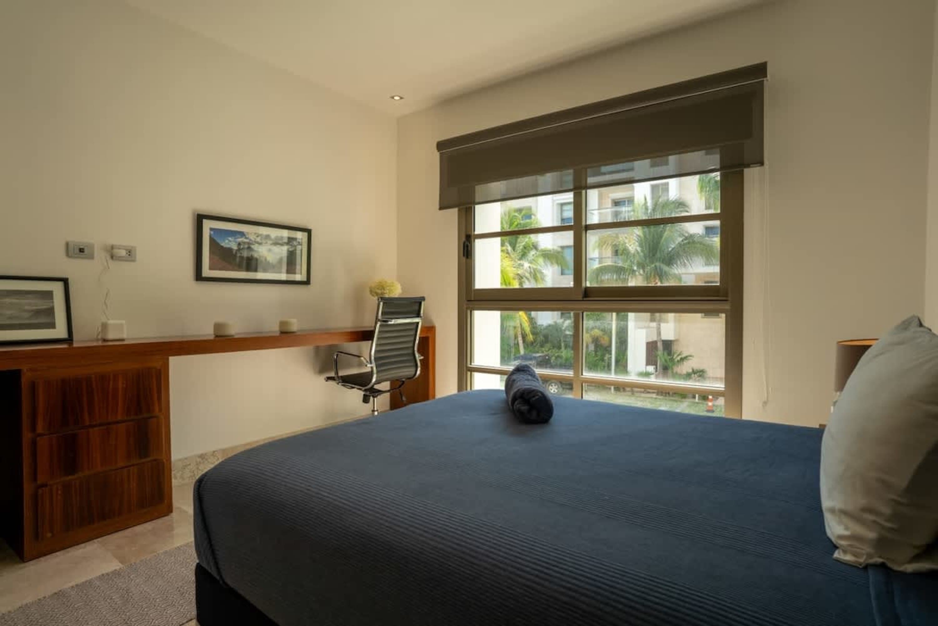 A bedroom with a desk and chair against the wall, and a large bed with a dark blue comforter.