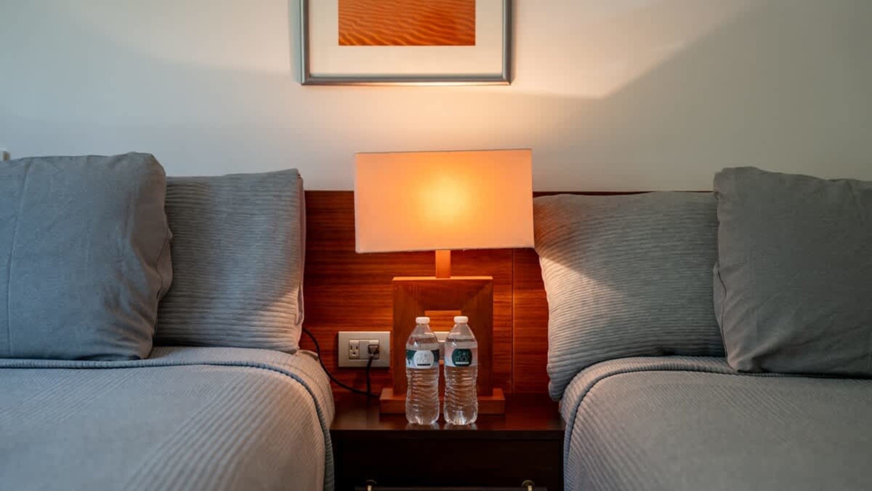 Two beds with gray pillows and a lamp between them.