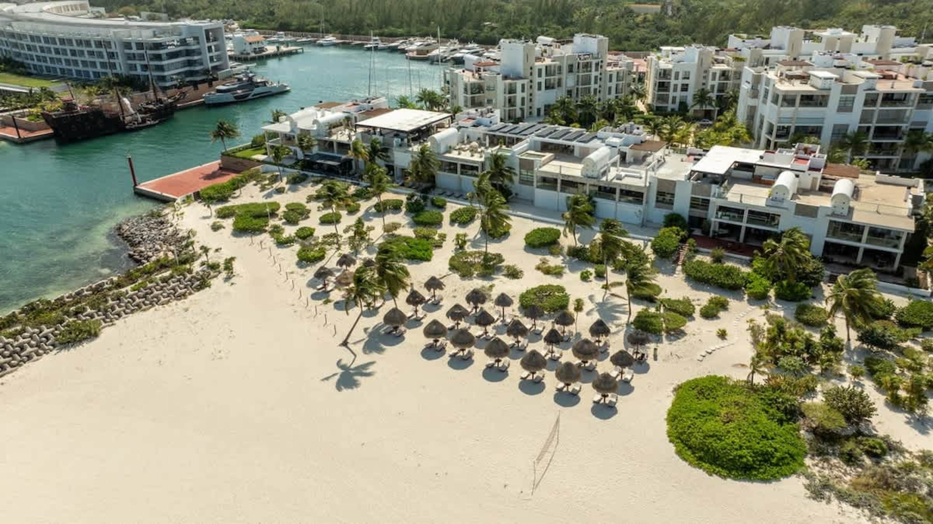 Aerial view of a tropical resort beach with white sand, palm trees, thatched umbrellas, and modern w