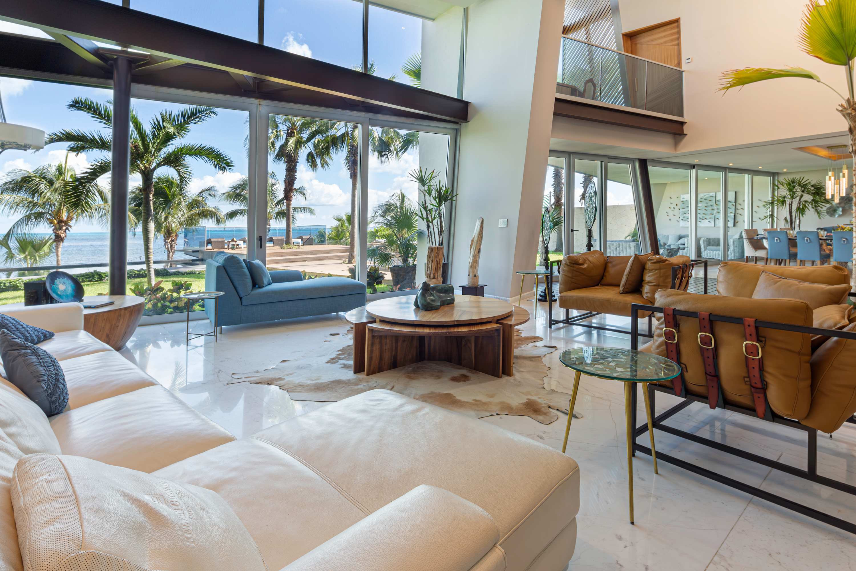 A modern living room with white leather sofas, tan leather chairs, and large windows overlooking the