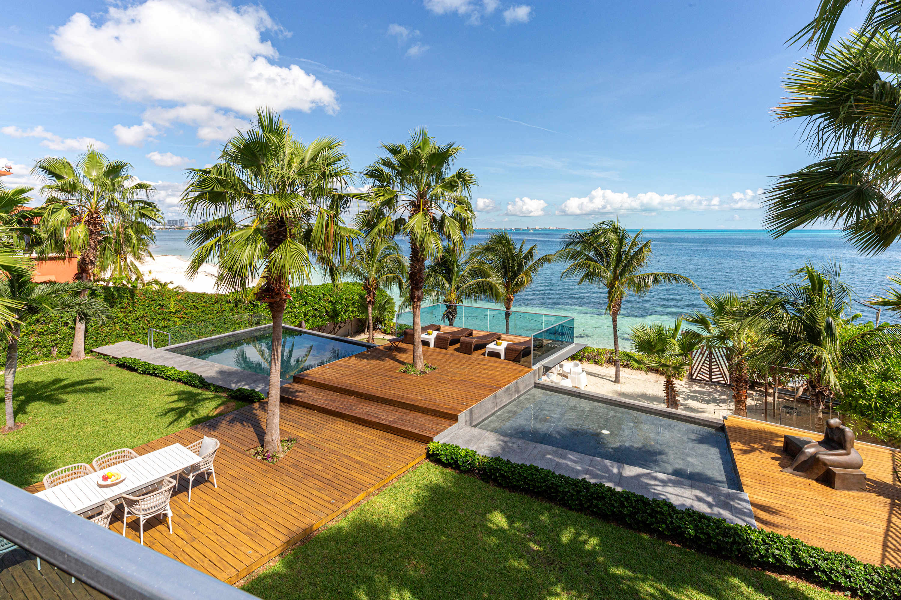 Luxury beachfront villa with multiple pools, wooden decks, palm trees, and ocean views.