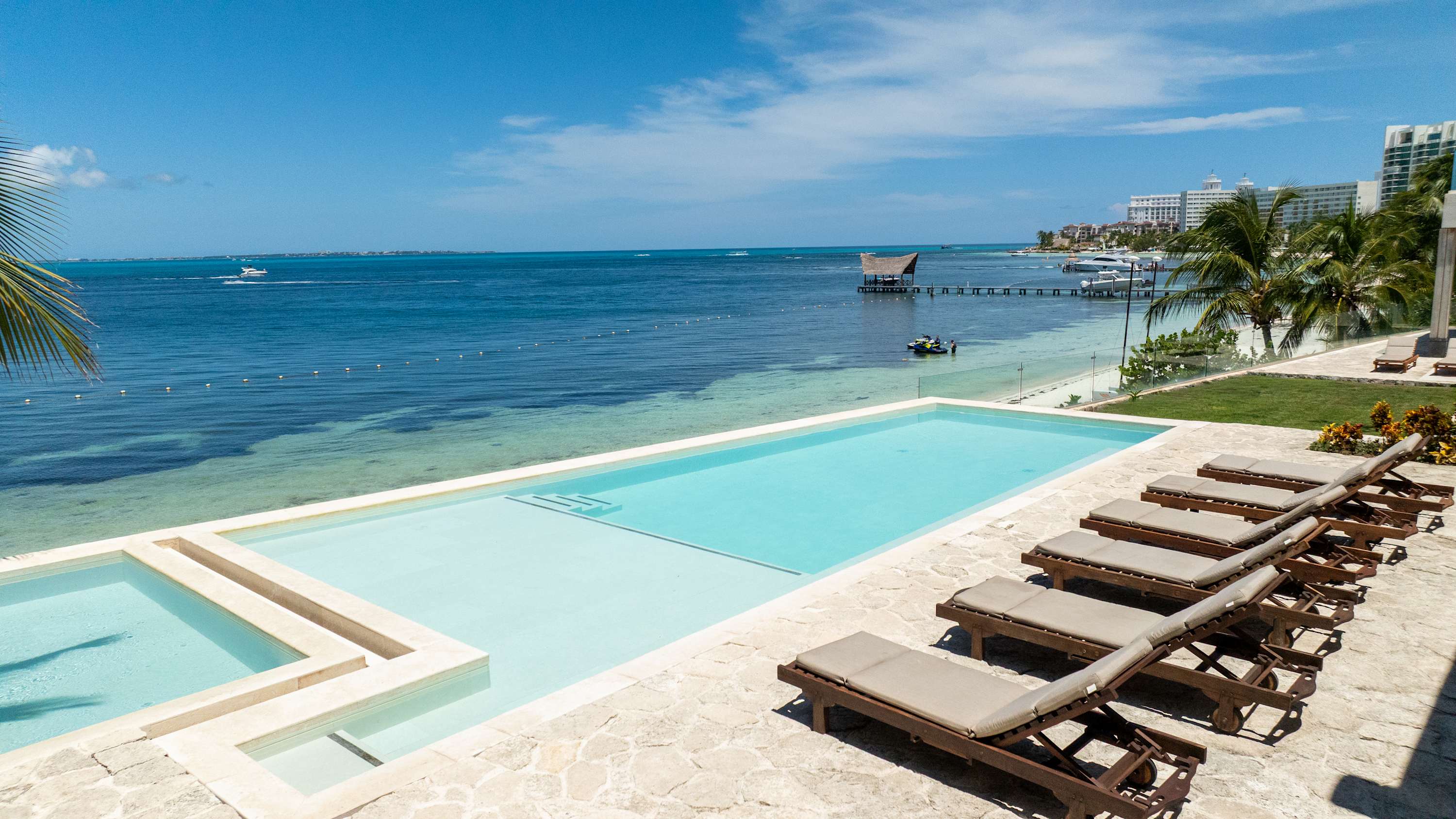 A resort pool with loungers overlooks a clear blue ocean with boats and buildings in the distance.