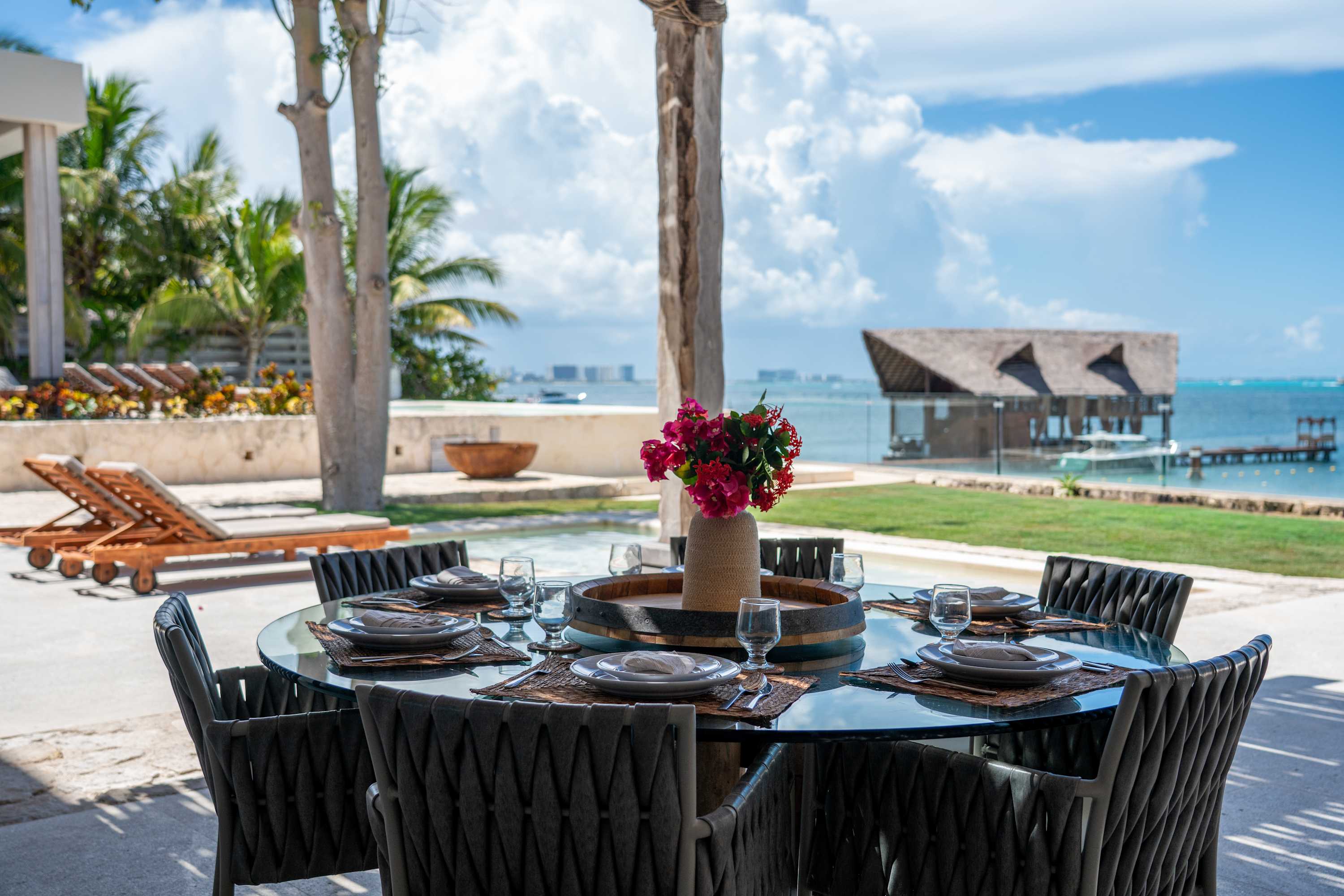An outdoor dining table set for six overlooks a tropical bay with a thatched-roof building.