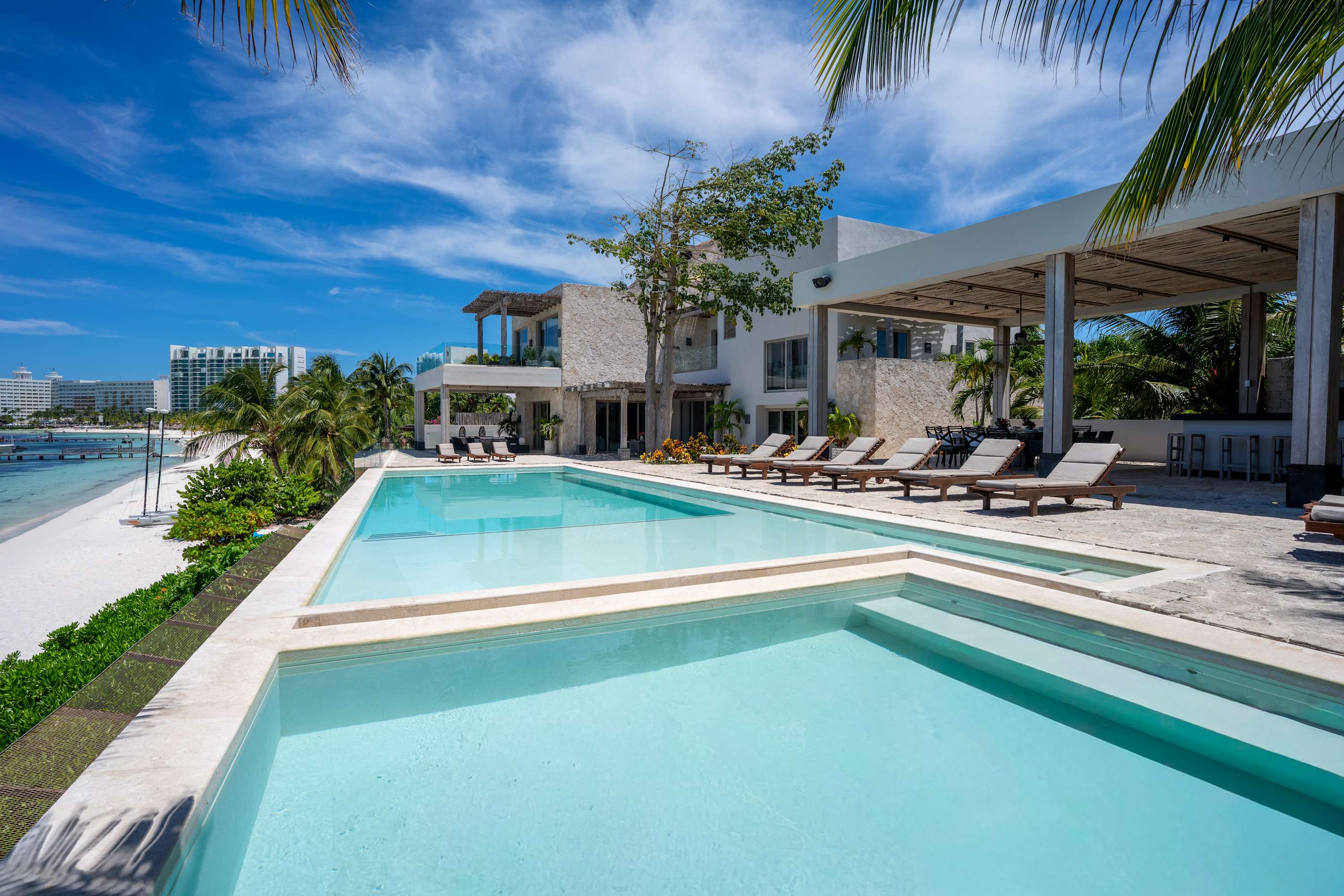 A luxury villa with a pool and lounge chairs overlooks a white sand beach and the ocean.