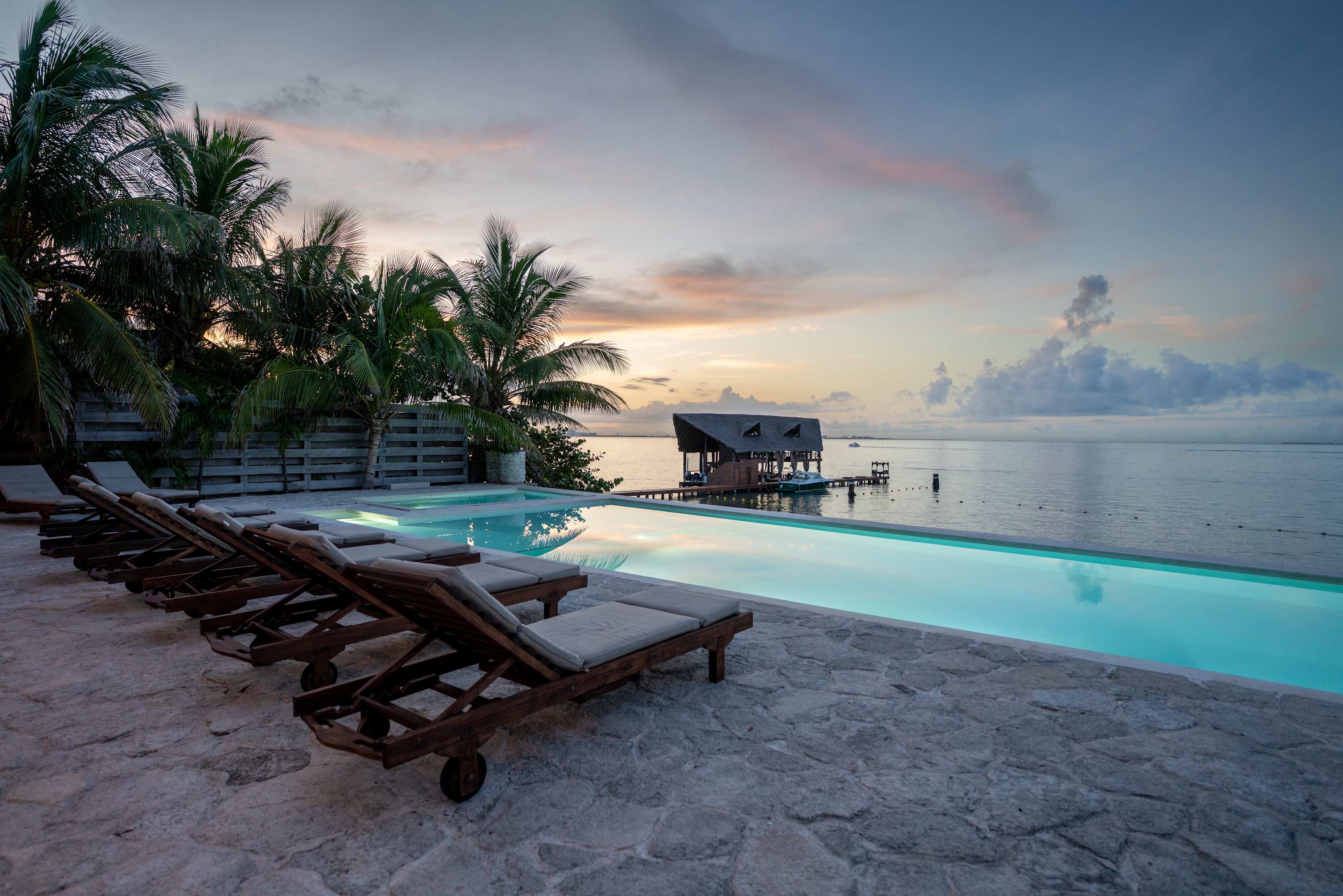 An infinity pool overlooks a calm ocean at sunset with palm trees and lounge chairs.