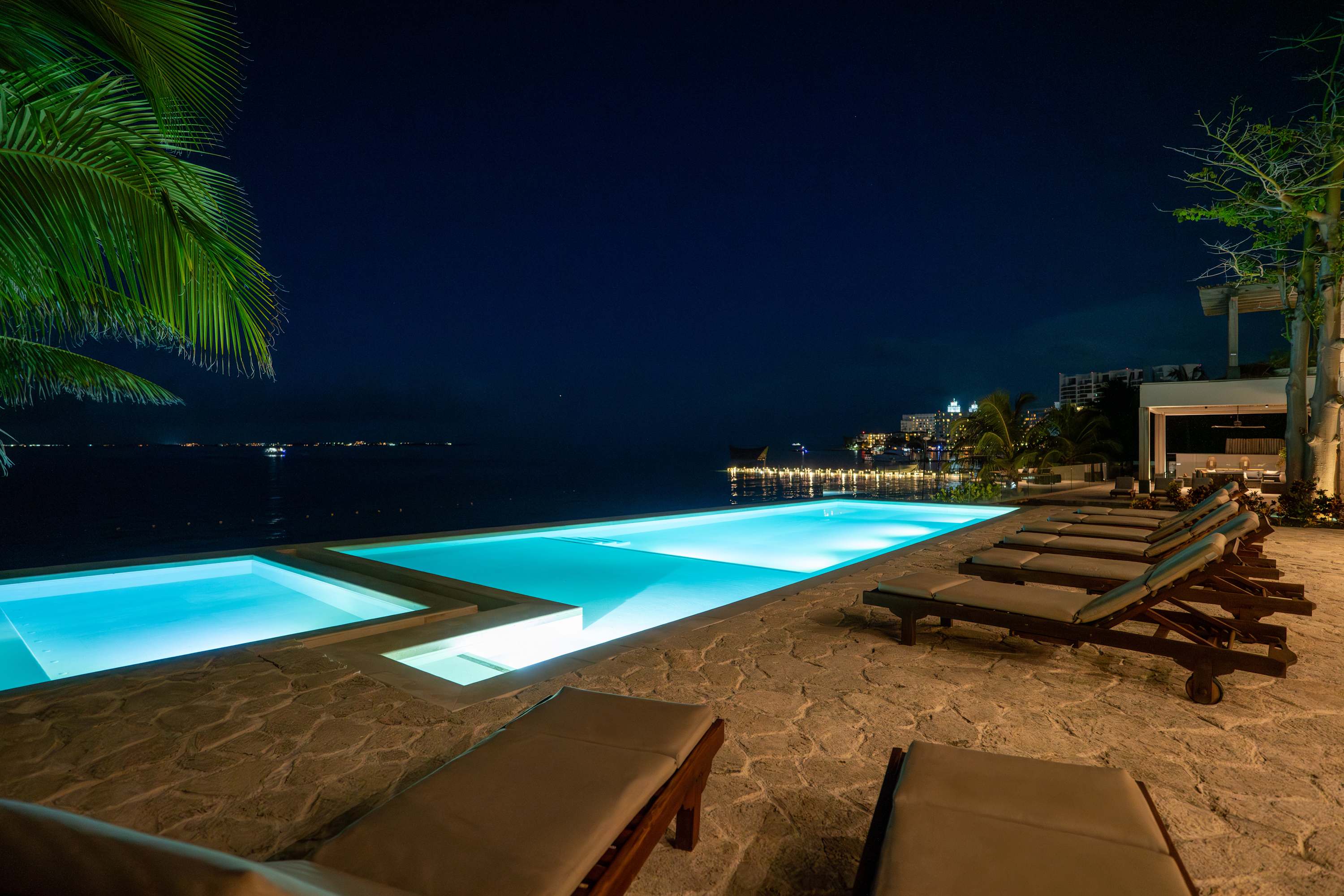 A night view of an infinity pool with lights, deck chairs and palm trees.