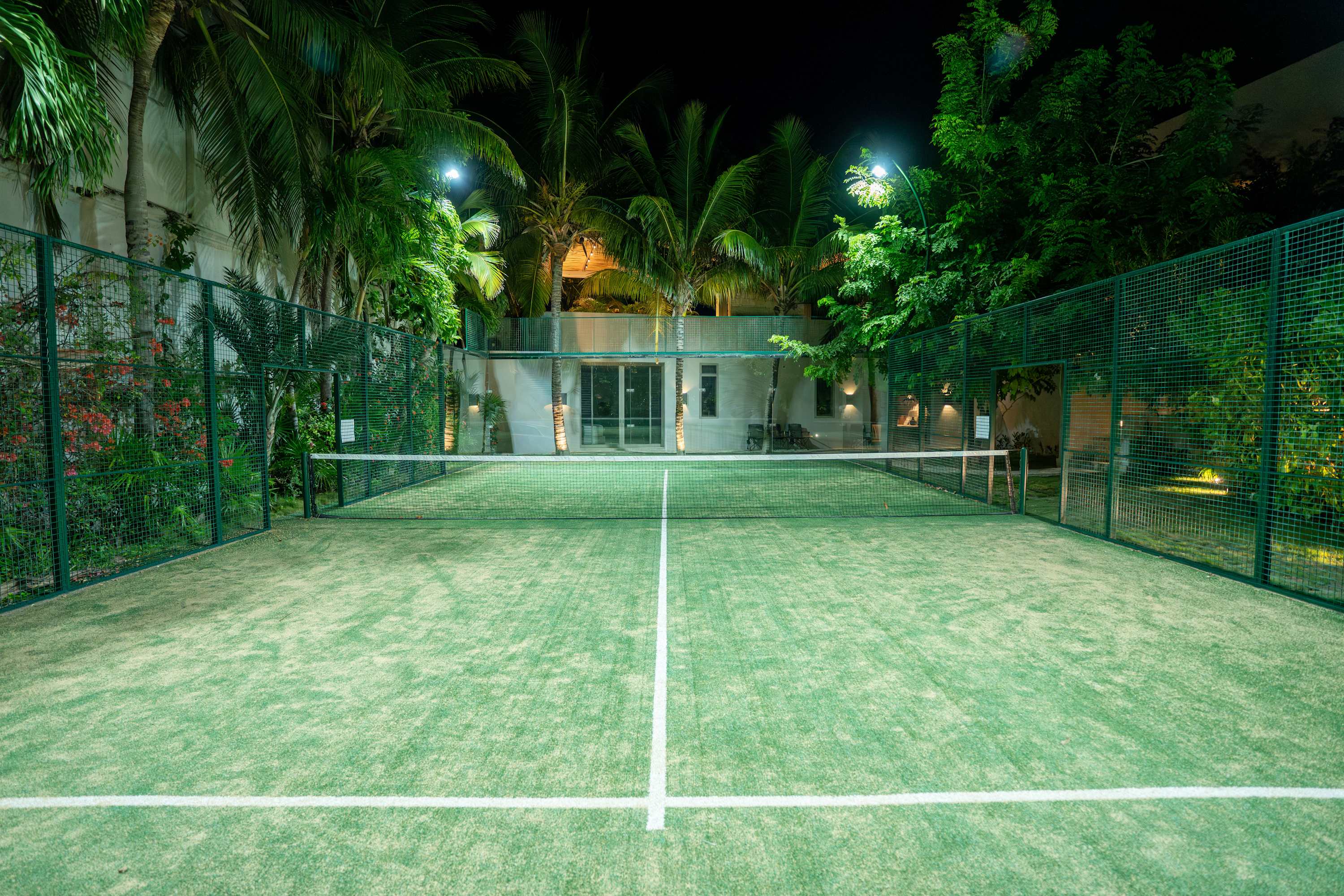 A night view of a padel court surrounded by palm trees and lush greenery, with a modern building in 