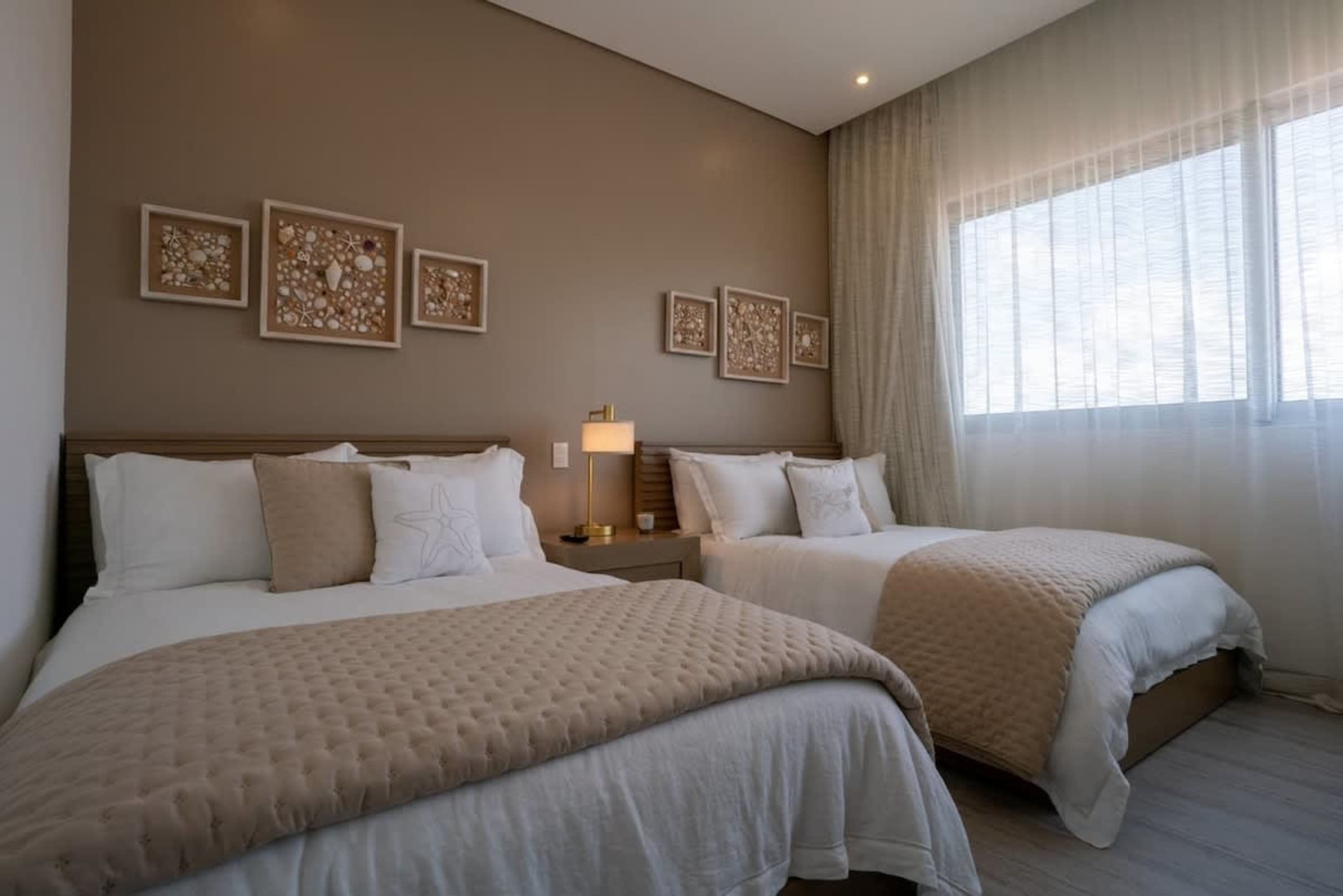 Two beds with white linens and tan quilted blankets in a room with seashell art and sheer curtains.