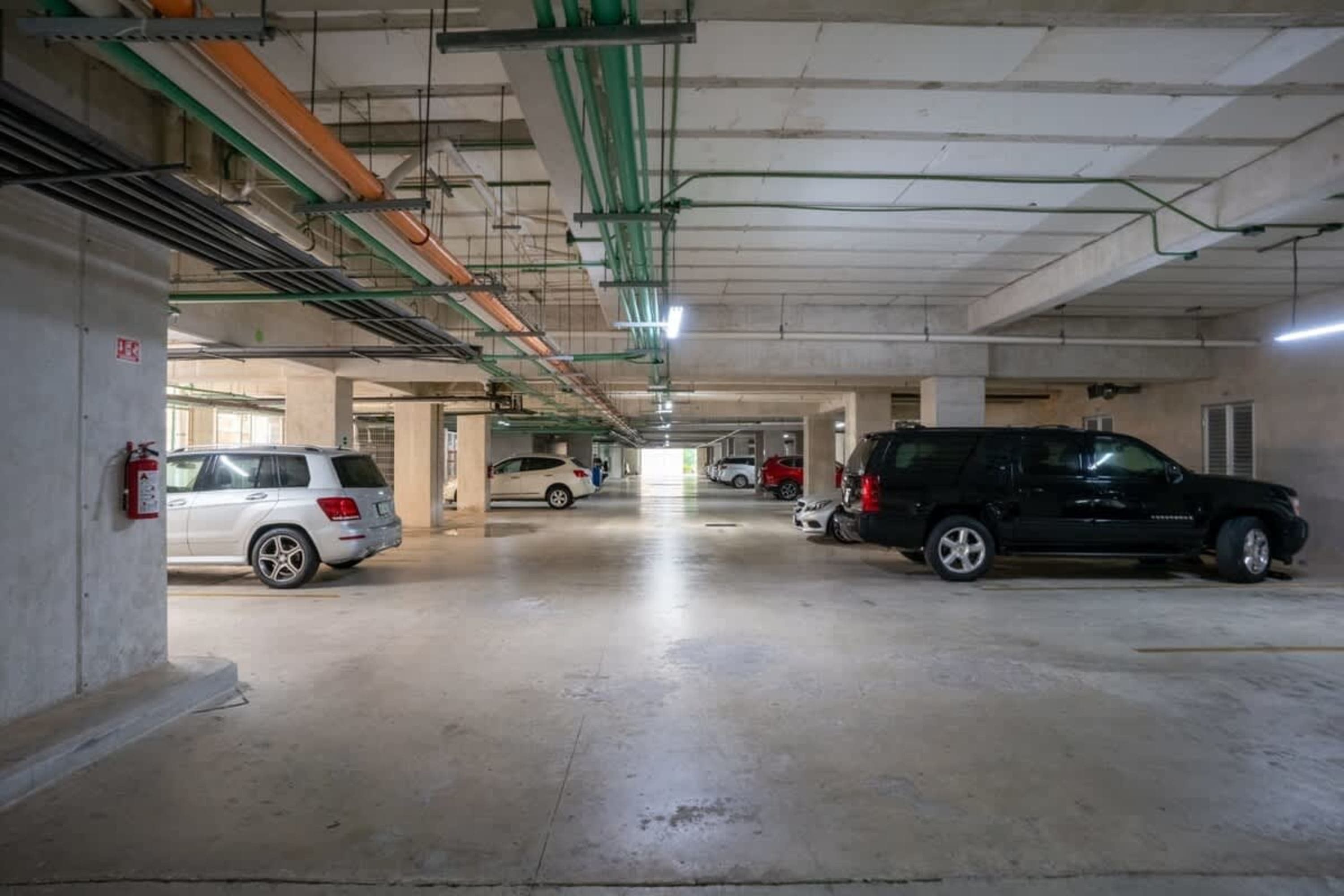 Cars parked in a well-lit underground parking garage with concrete pillars and pipes overhead.