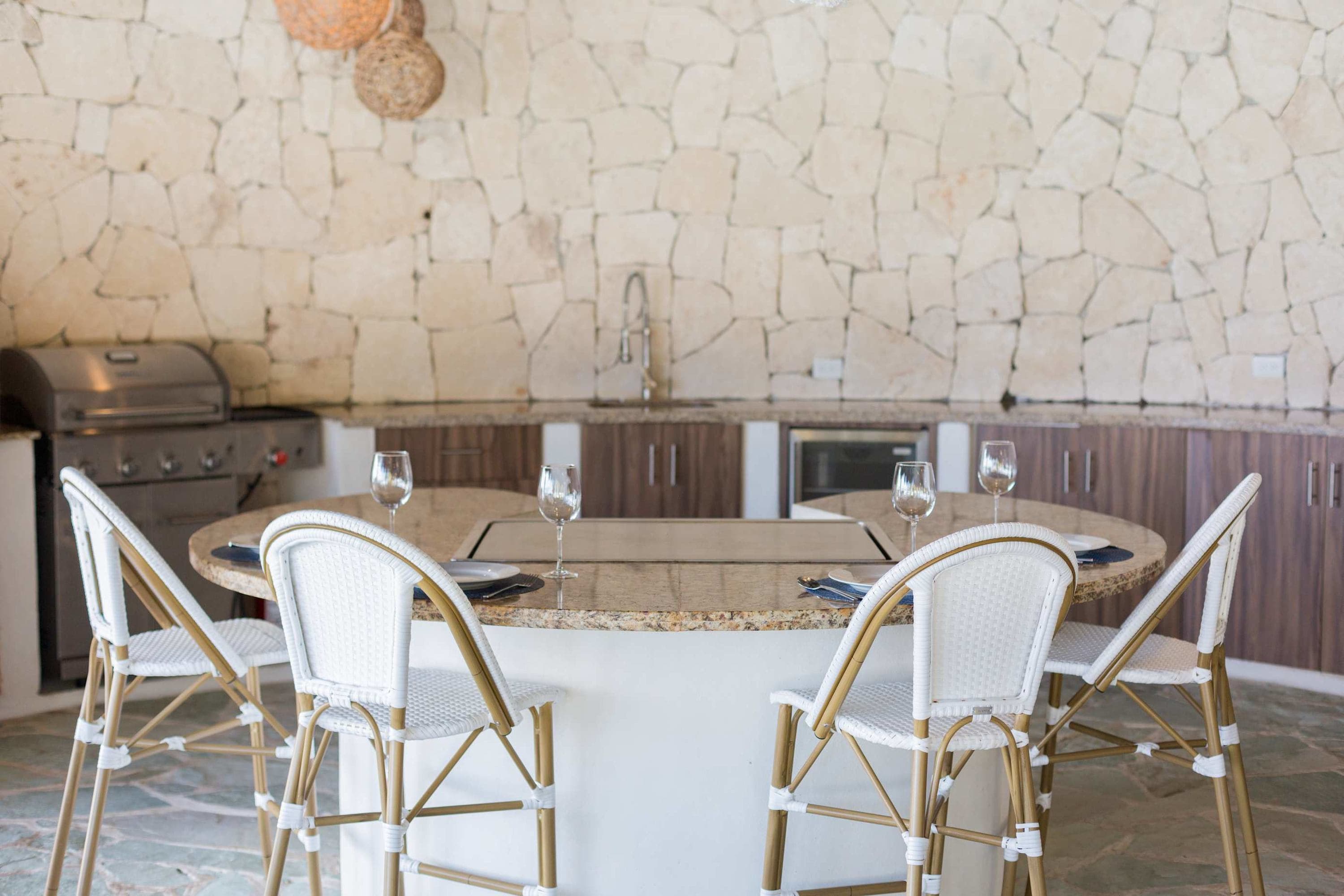 An outdoor kitchen with a grill and bar seating area features a stone wall and granite countertops.