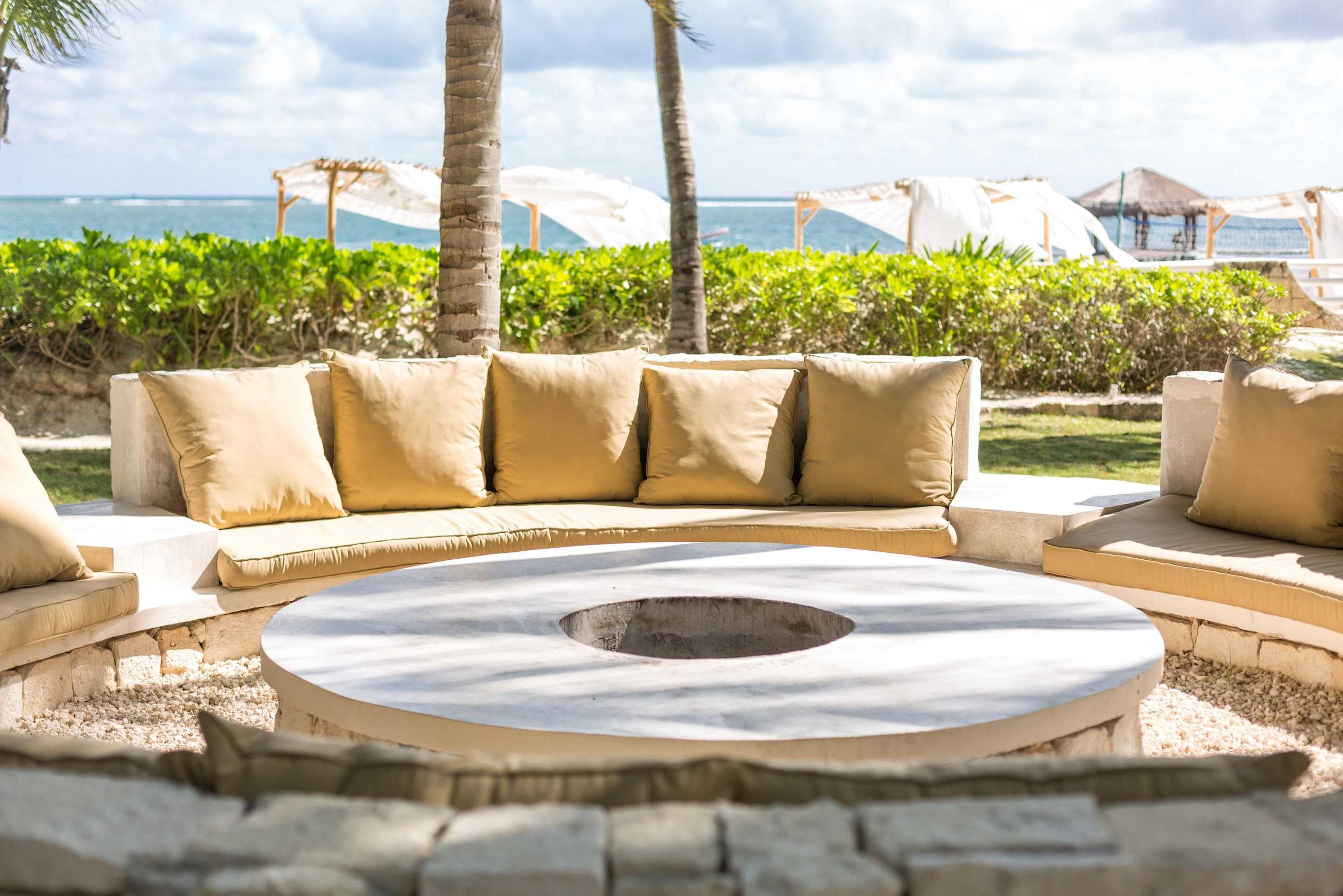 A beachside seating area with a U-shaped sofa and fire pit, overlooking the ocean.