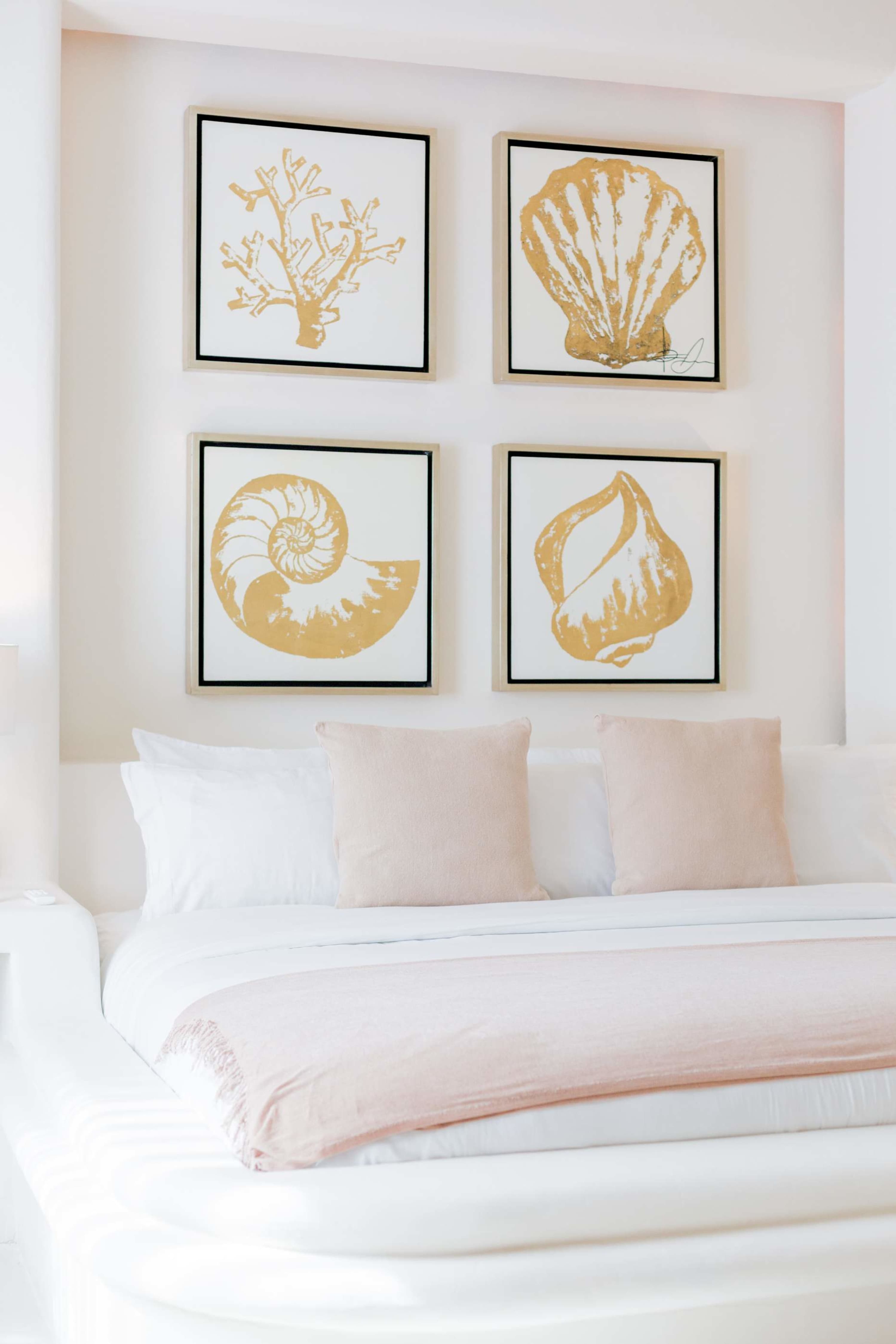 Four framed seashell and coral artworks hang above a bed with white linens and pink accents.