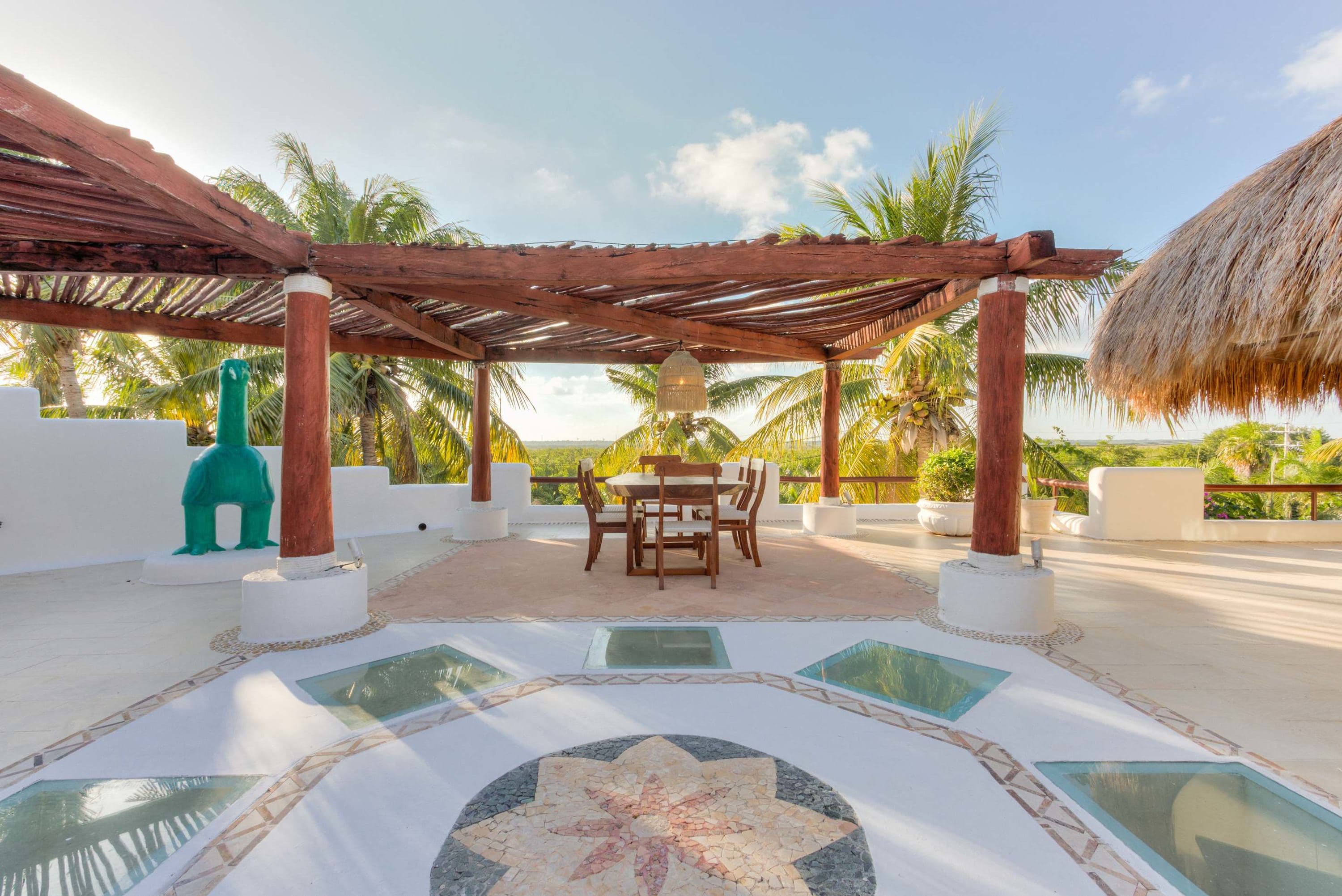 A rooftop patio with a pergola, thatched roof, dining set, and a turquoise bird sculpture.