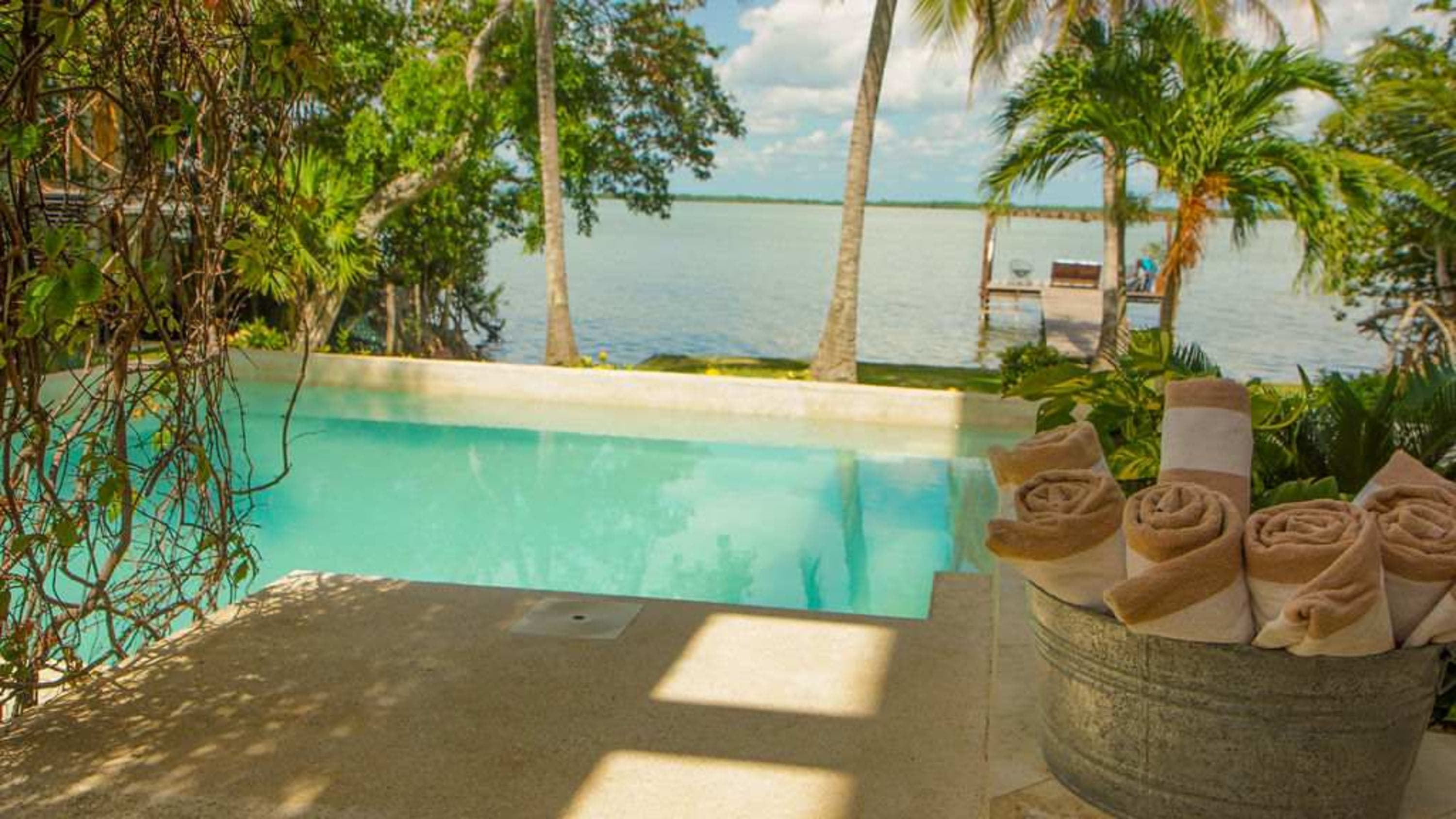A tropical poolside with towels in a galvanized bucket, overlooking a serene lake.