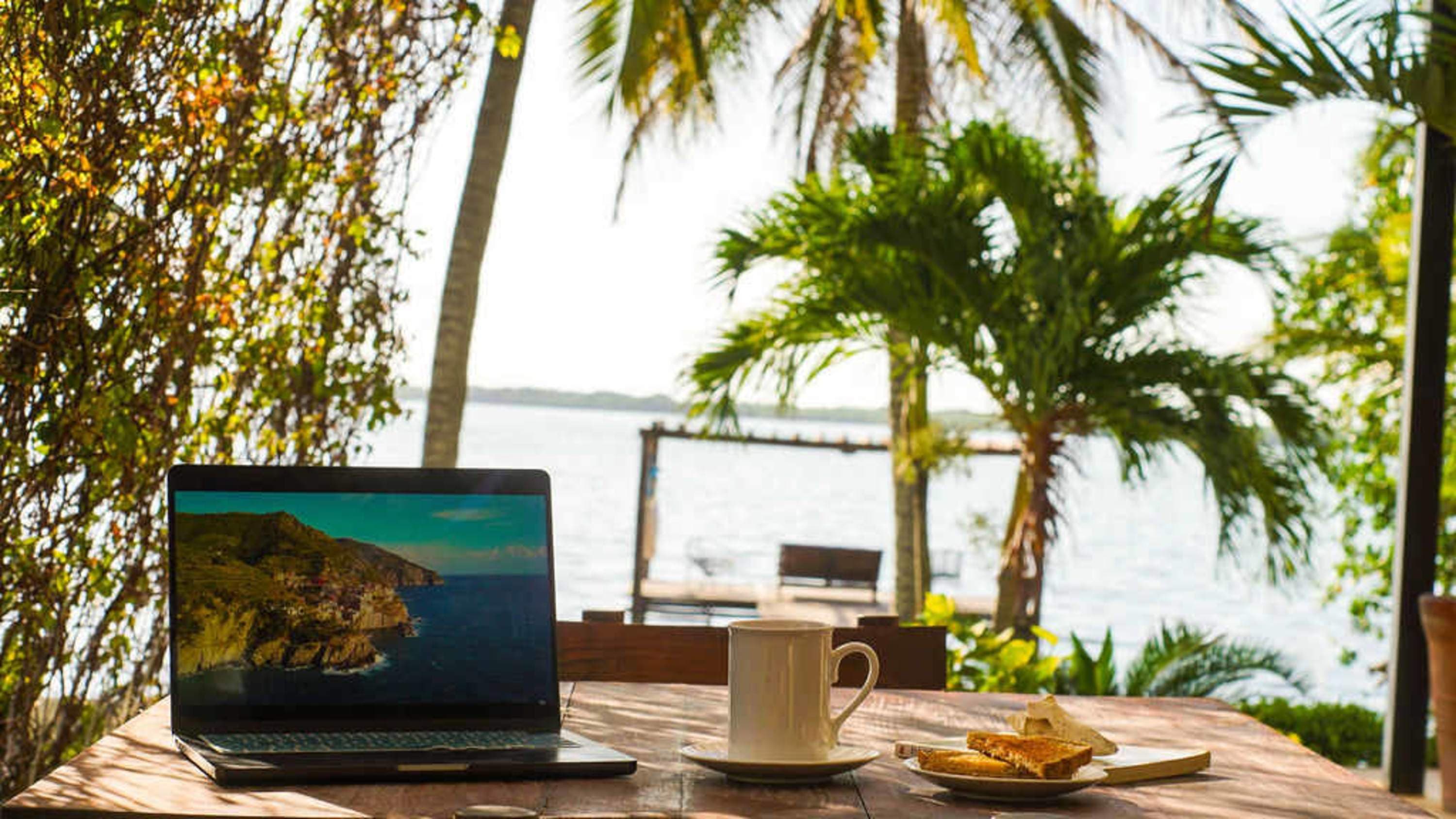 Laptop, coffee, and toast on a table with a view of palm trees and the ocean.