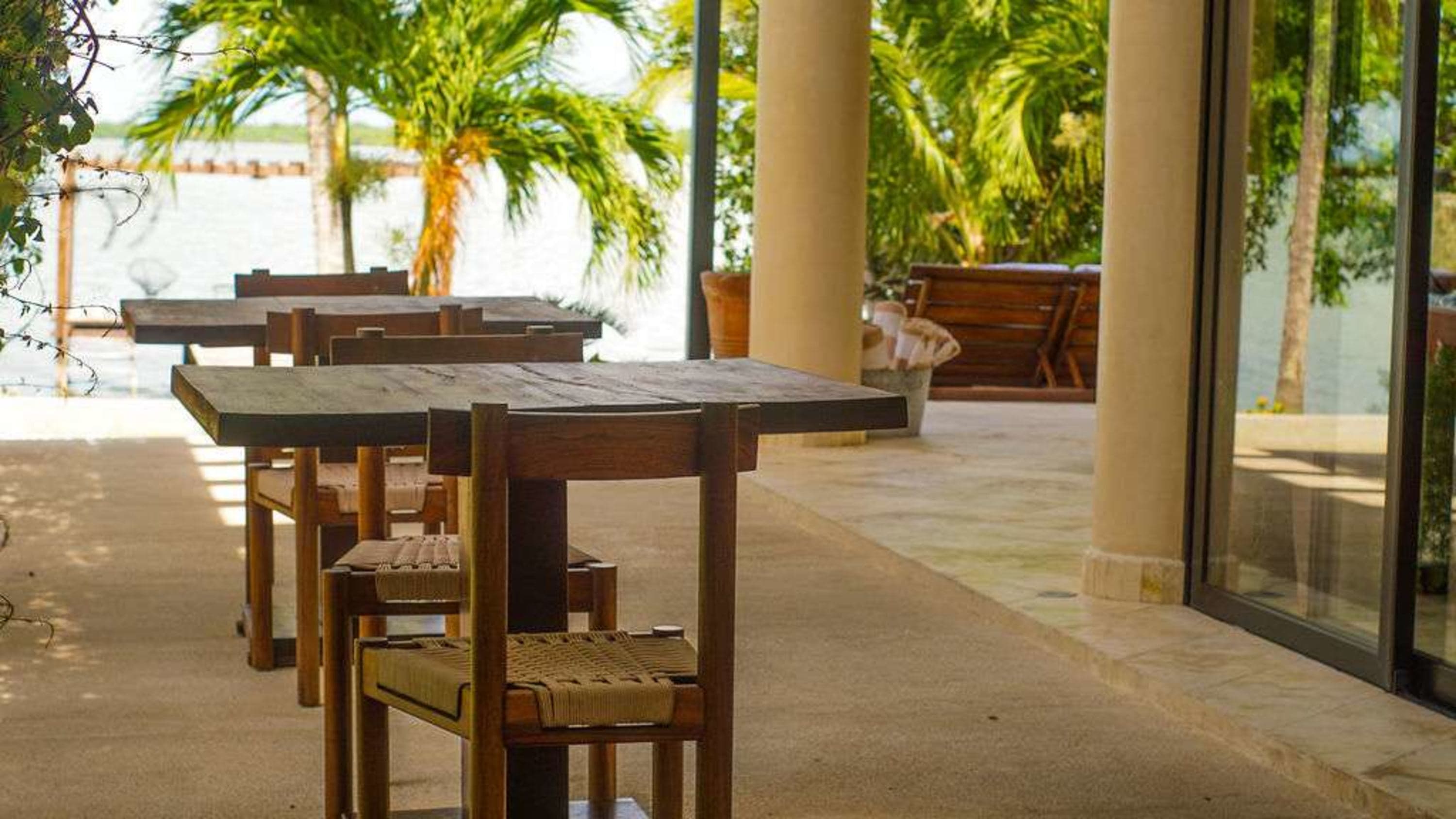 Outdoor seating area with tables and chairs, palm trees and water in the background.
