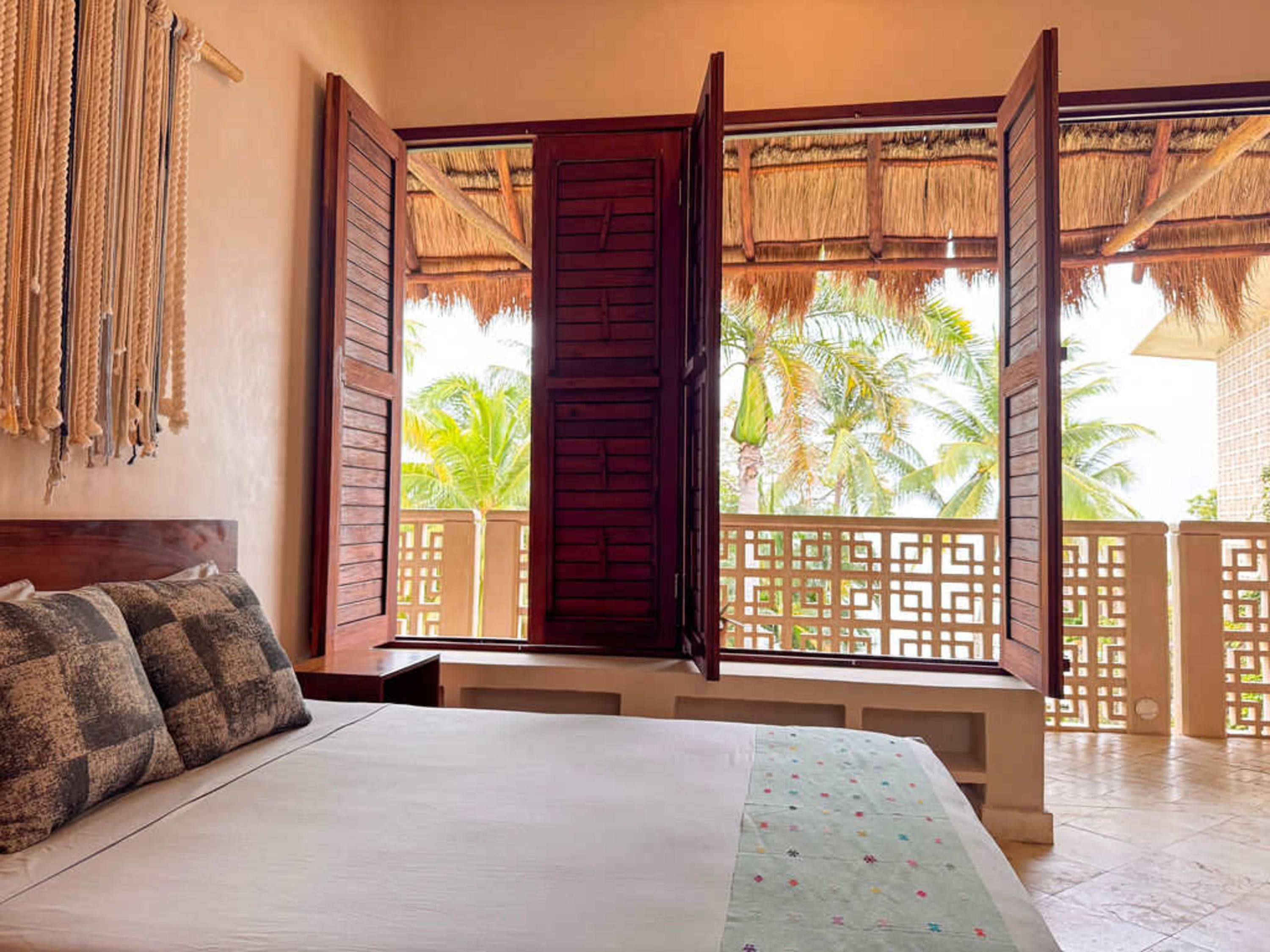 Bedroom with open wooden shutters revealing a tropical balcony with palm trees.
