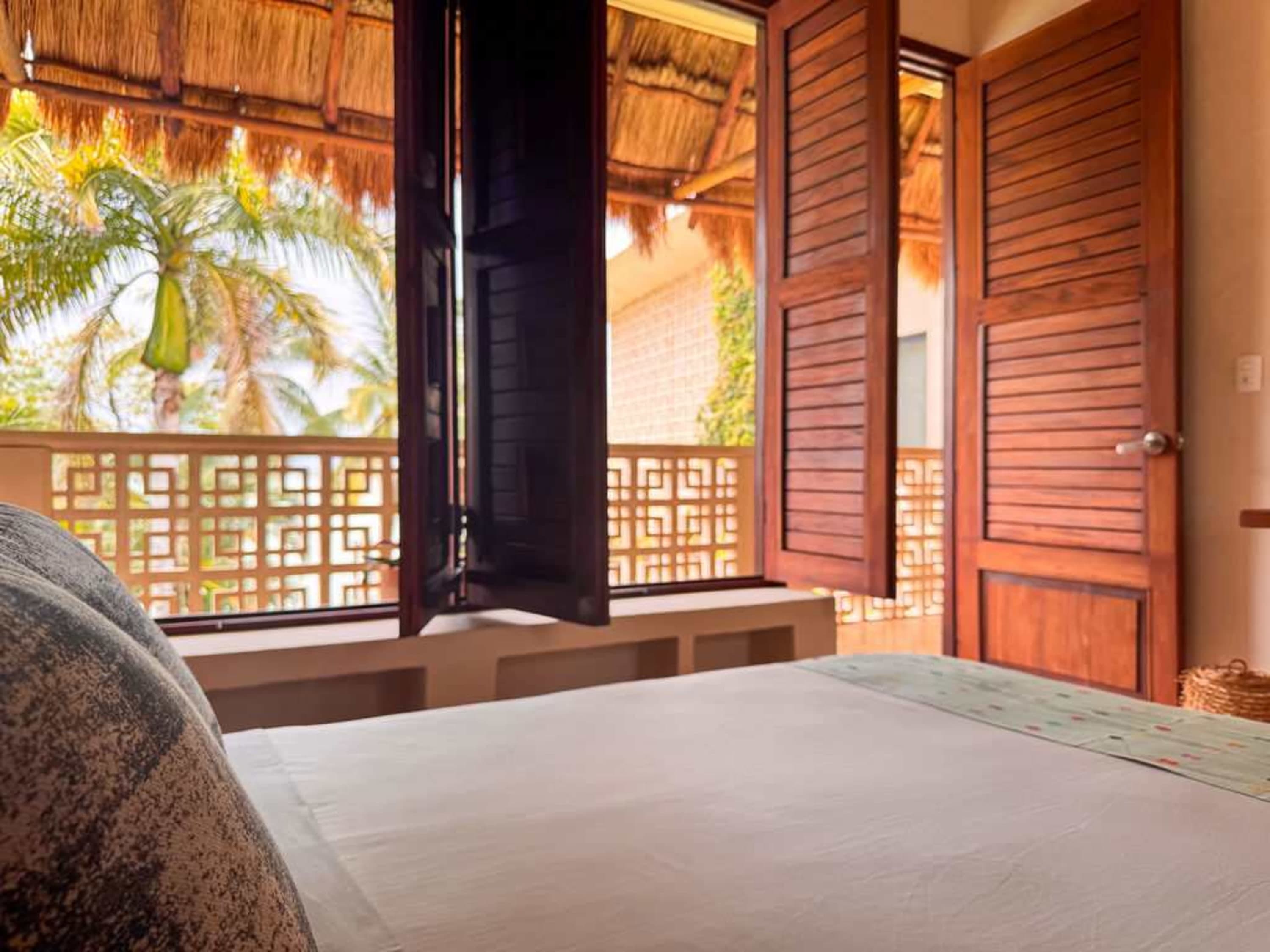 Bedroom with a view of palm trees through open wooden shutters, a bed in the foreground.