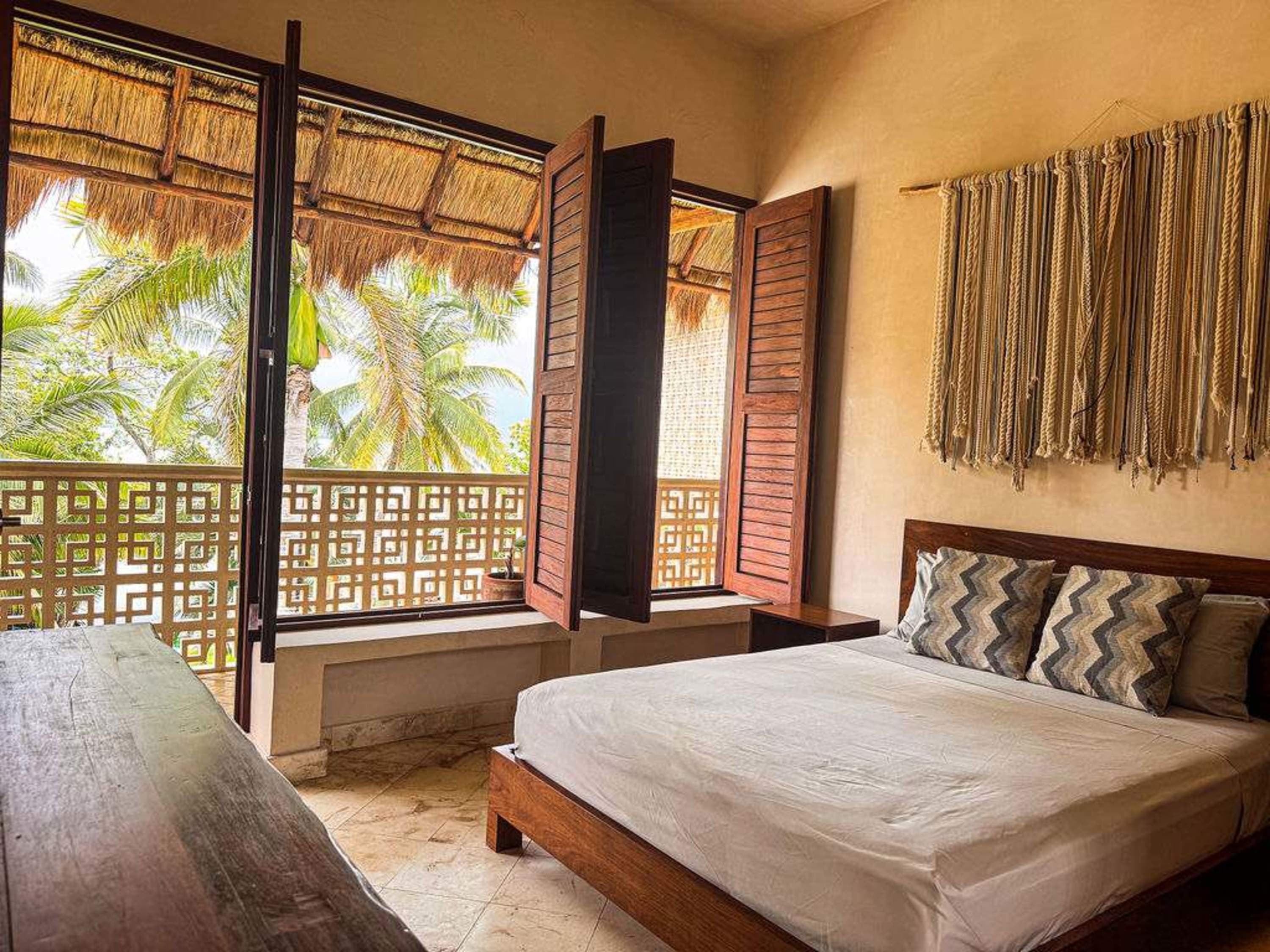 A bed with pillows, a wooden bench, and open windows with a view of palm trees.