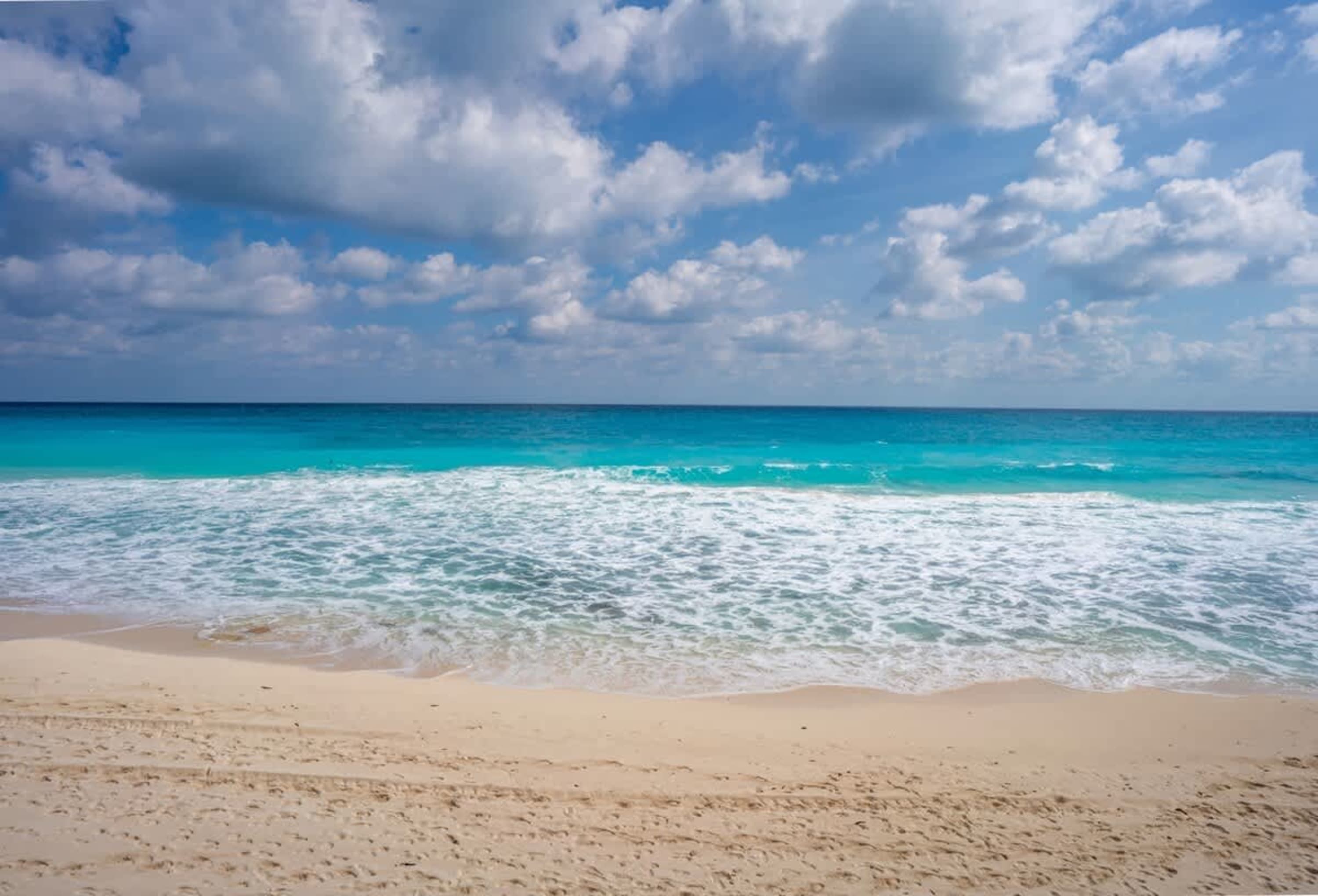 A sandy beach meets the turquoise ocean under a cloudy blue sky.