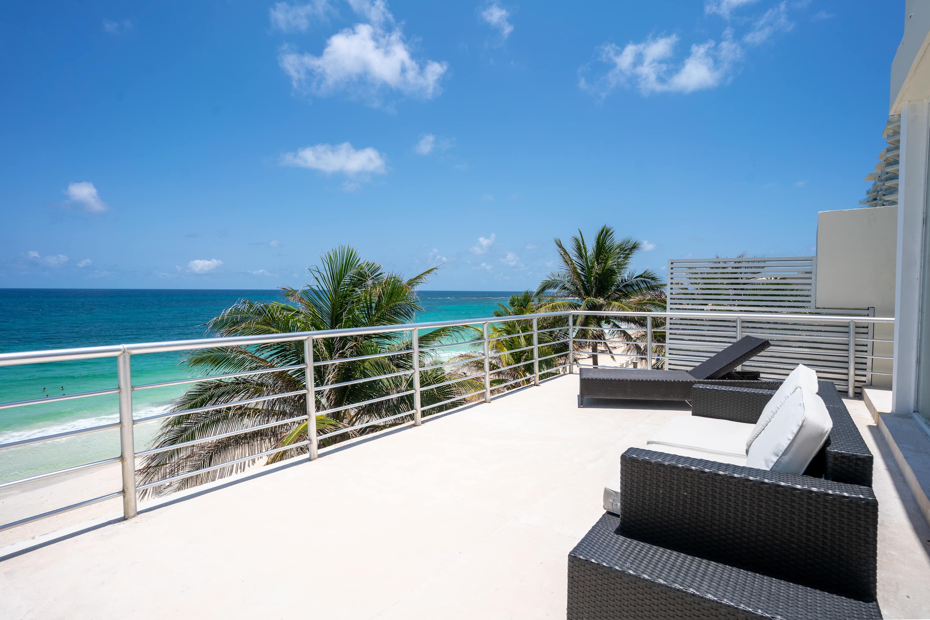 A balcony with lounge chairs overlooks the ocean and palm trees under a blue sky.
