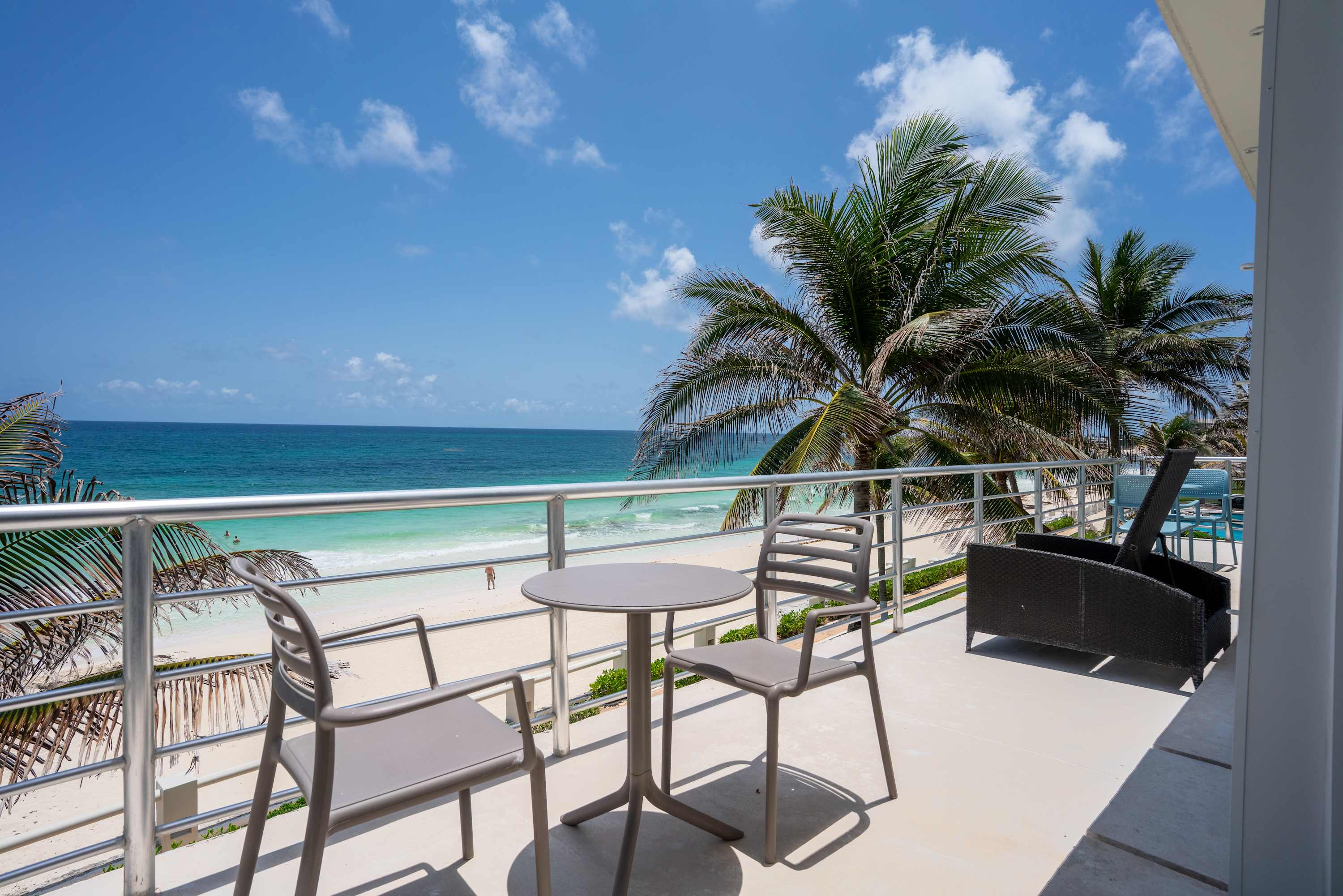 Balcony view of a tropical beach with palm trees, ocean, and a table with chairs.