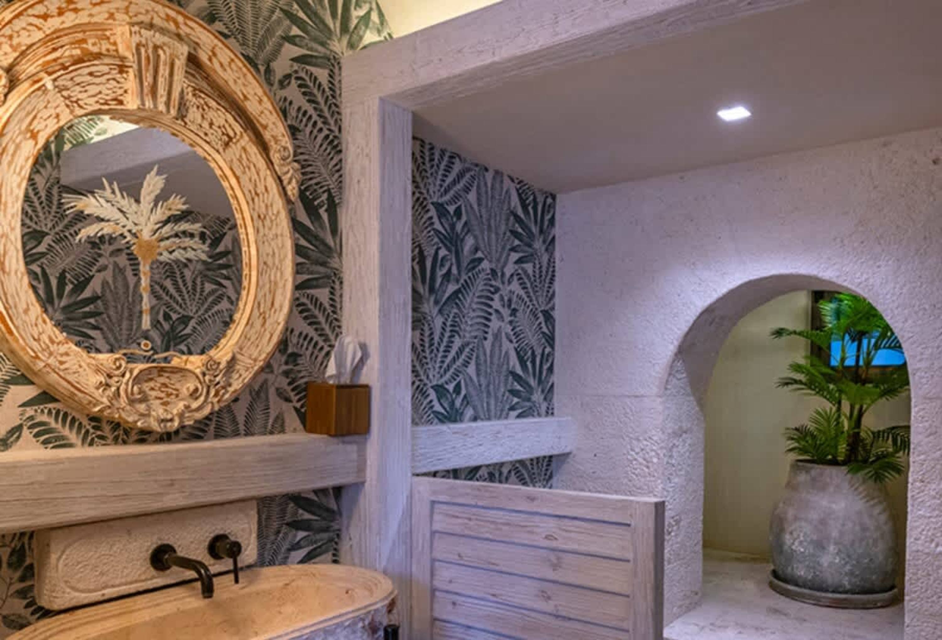 A bathroom with a palm leaf wallpaper, a rustic mirror and sink, and an arched doorway leading to a 