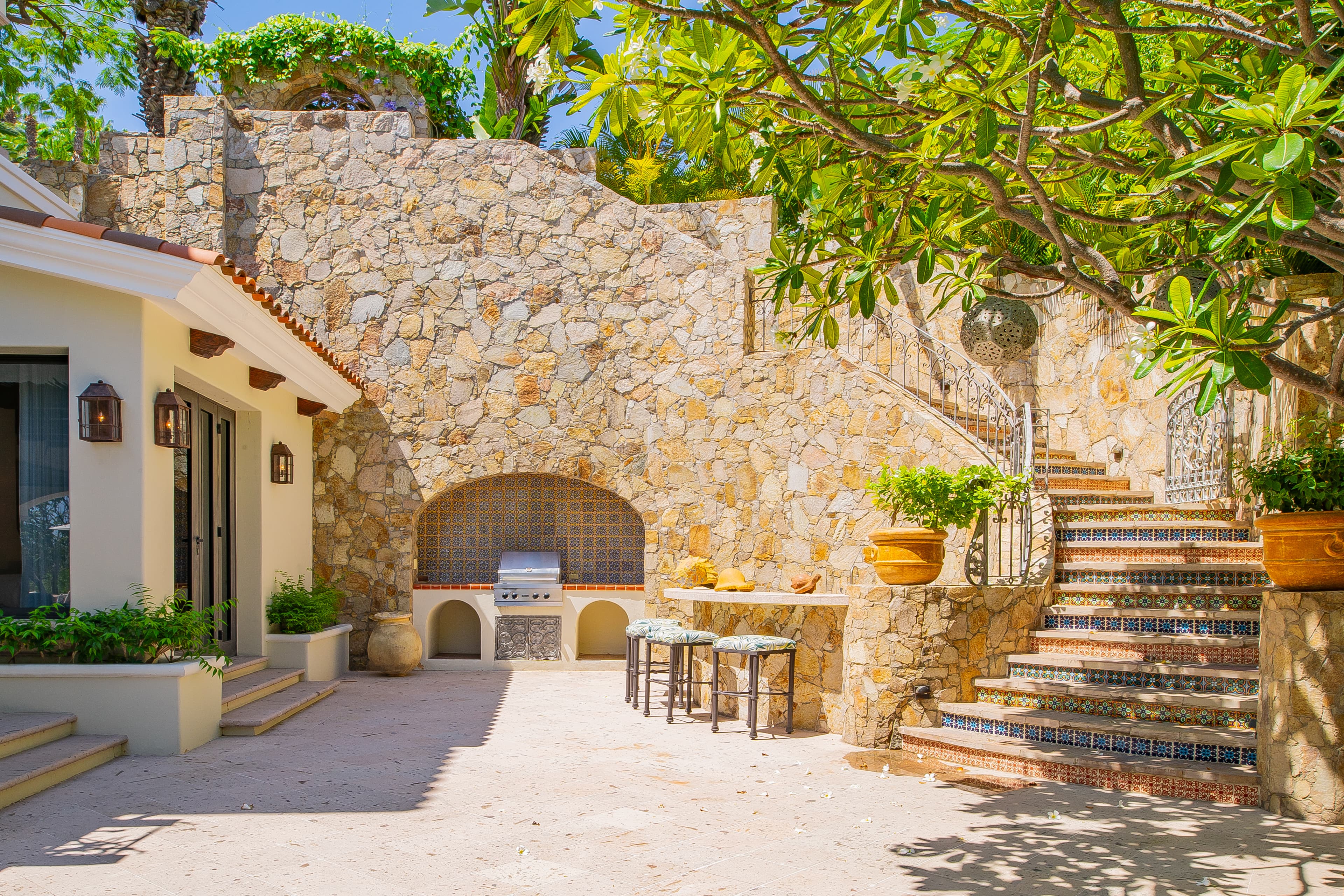 A sunlit courtyard features beautiful stone walls, a charming built-in grill, and a tiled mosaic staircase winding upward beneath lush tropical foliage—a perfect spot to unwind.
