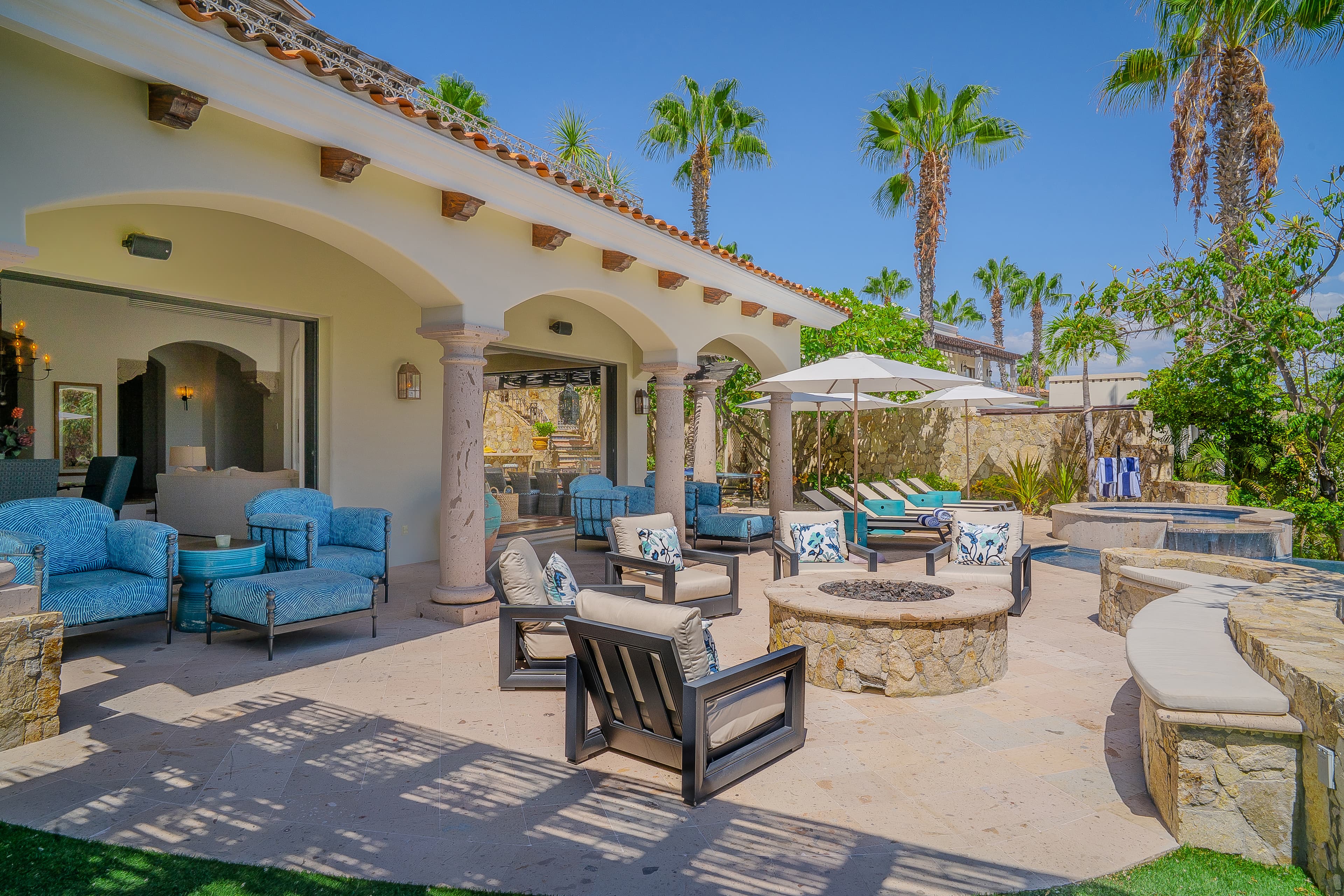 Relax in your private outdoor oasis featuring elegant archways, plush seating, a fire pit, and poolside loungers surrounded by swaying palms and lush greenery.