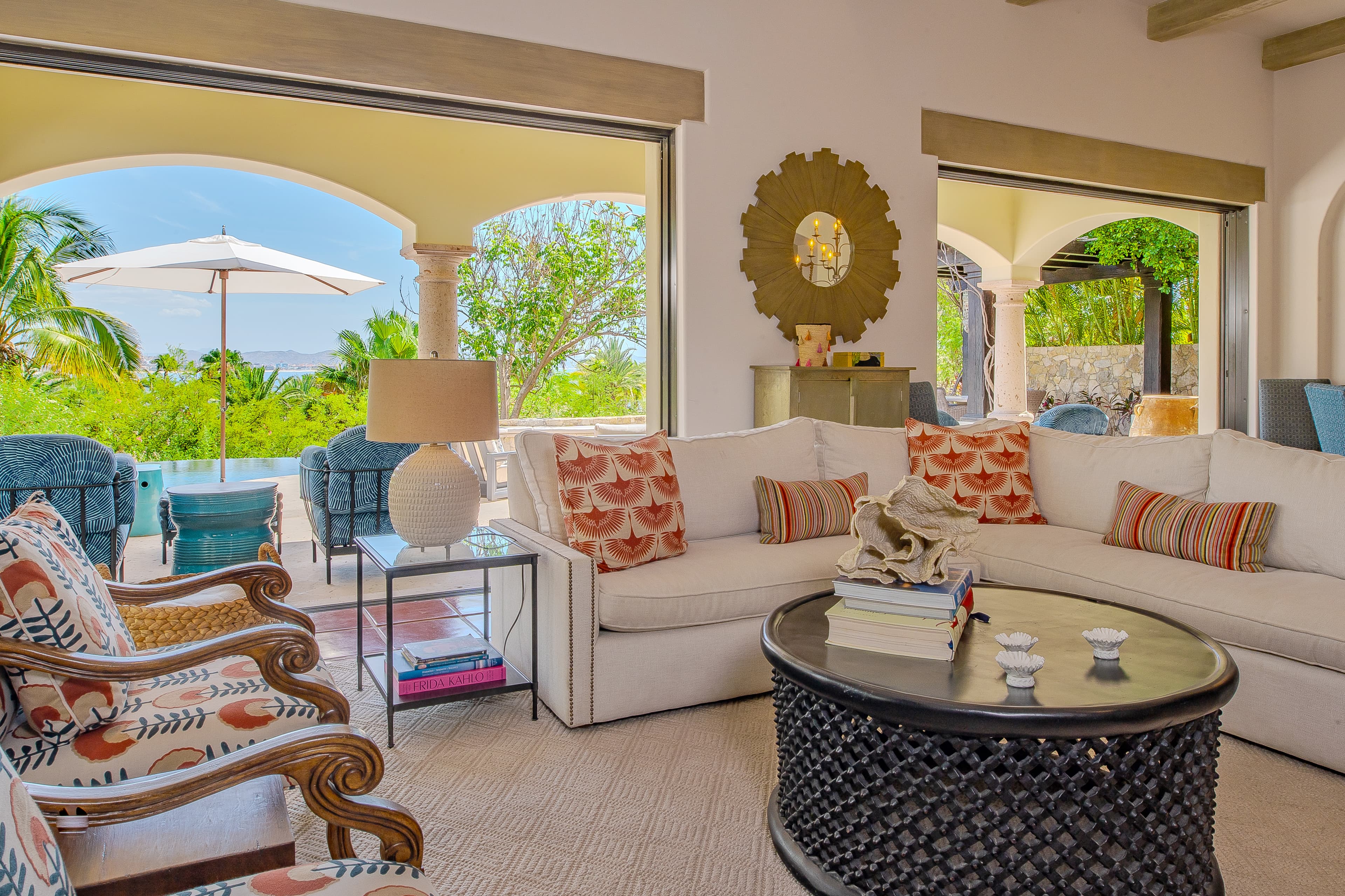 Step into your serene living space where plush seating and vibrant tropical décor invite you to unwind, while open archways frame lush greenery and sparkling pool views beyond.