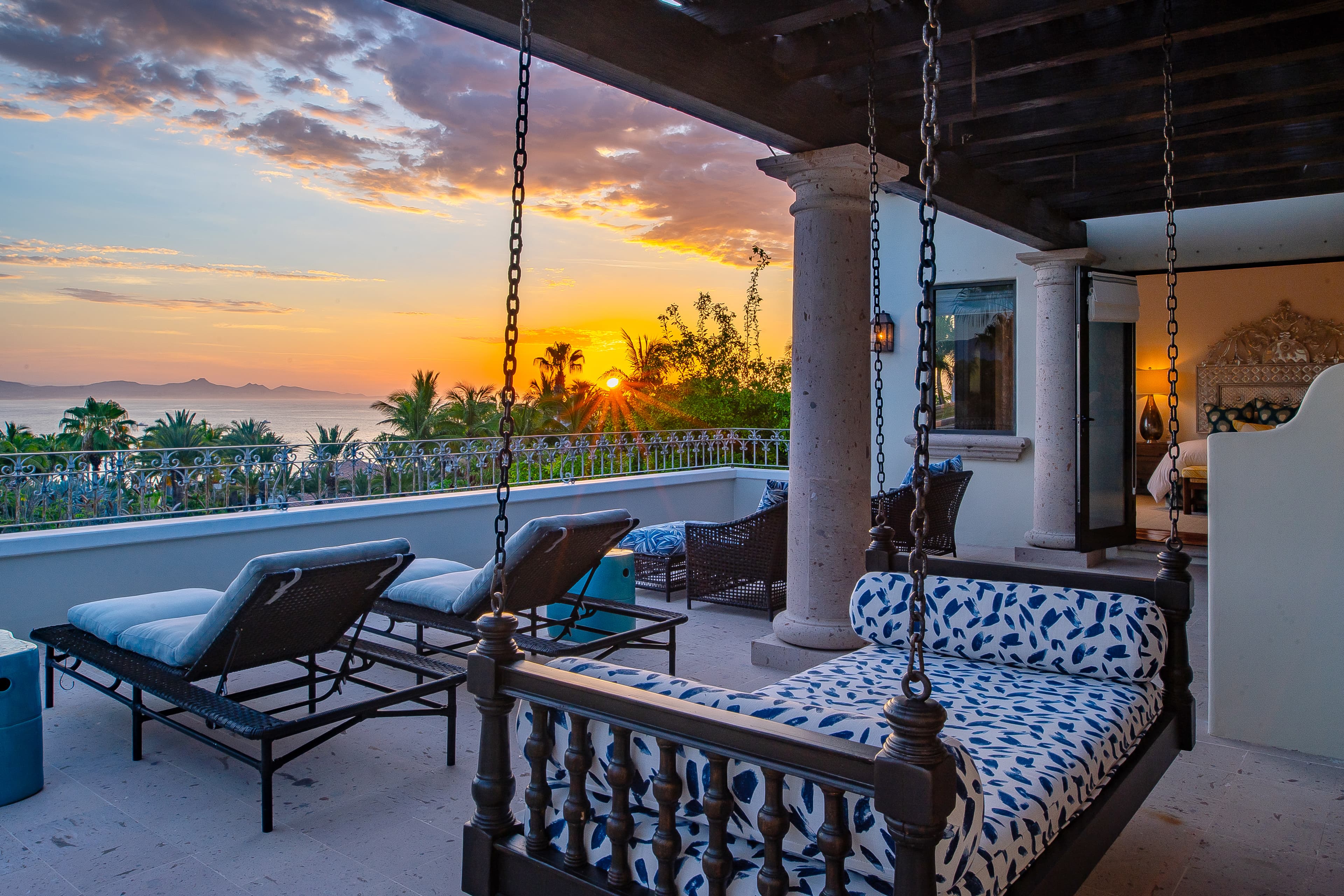 Elegant terrace with suspended swing, sun loungers, and colonial columns framing a stunning coastal sunset over tropical palms and distant islands.
