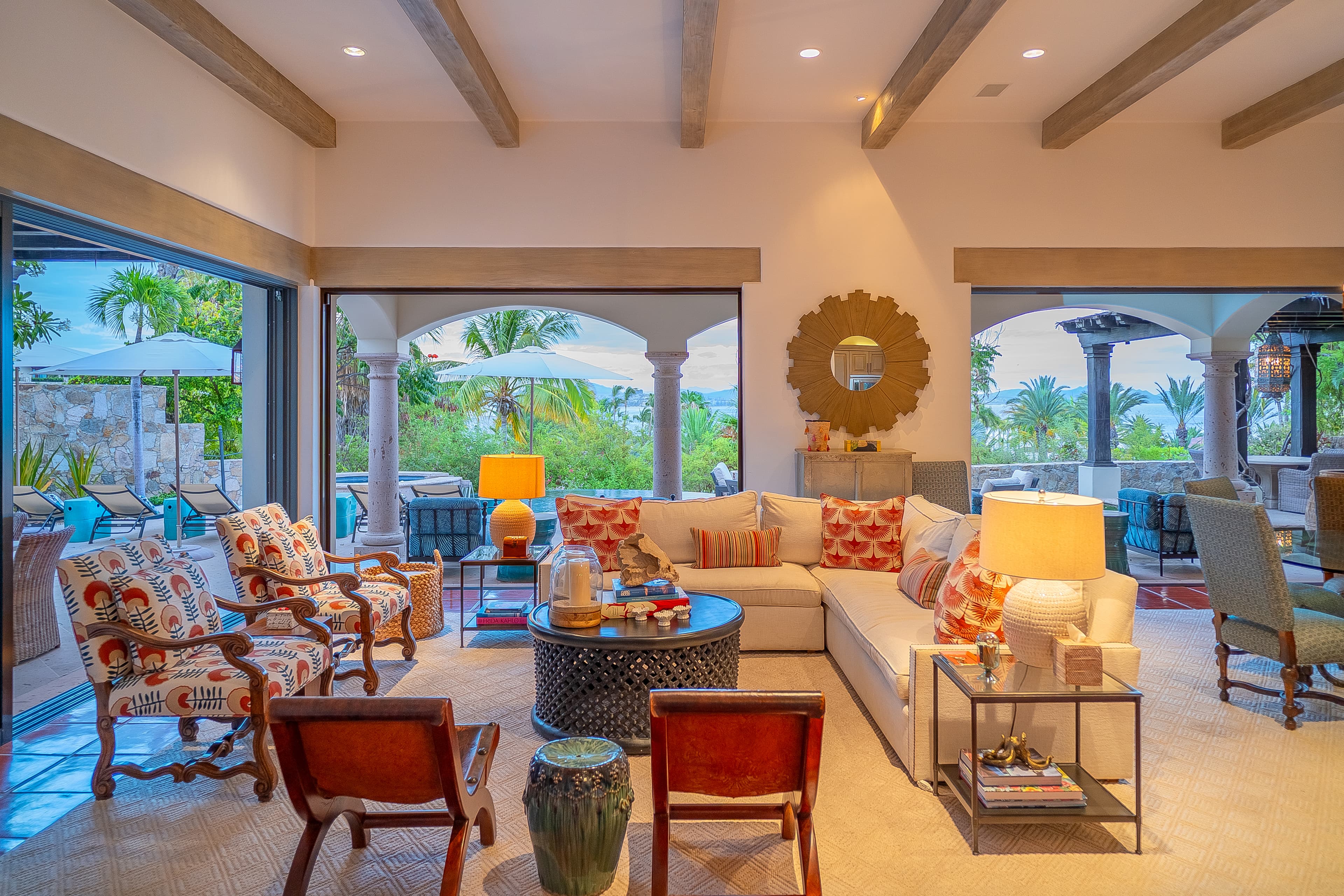 Sink into plush seating surrounded by vibrant tropical colors and natural light streaming through open arches. Your living space flows seamlessly to outdoor lounging and pool views, inviting relaxation under elegant beamed ceilings.