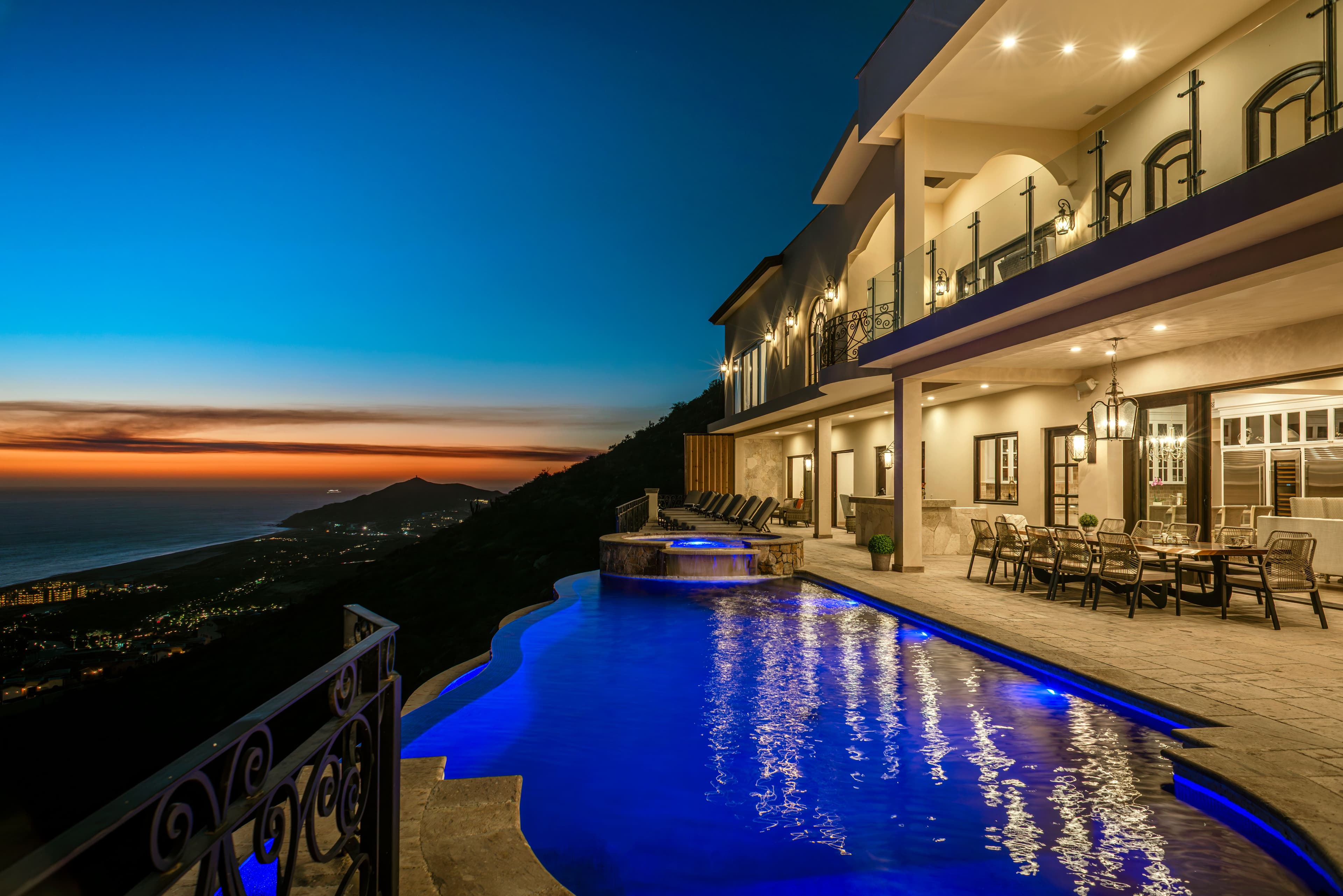 Luxurious hillside villa featuring illuminated pool and panoramic sunset views over mountainous coastline.