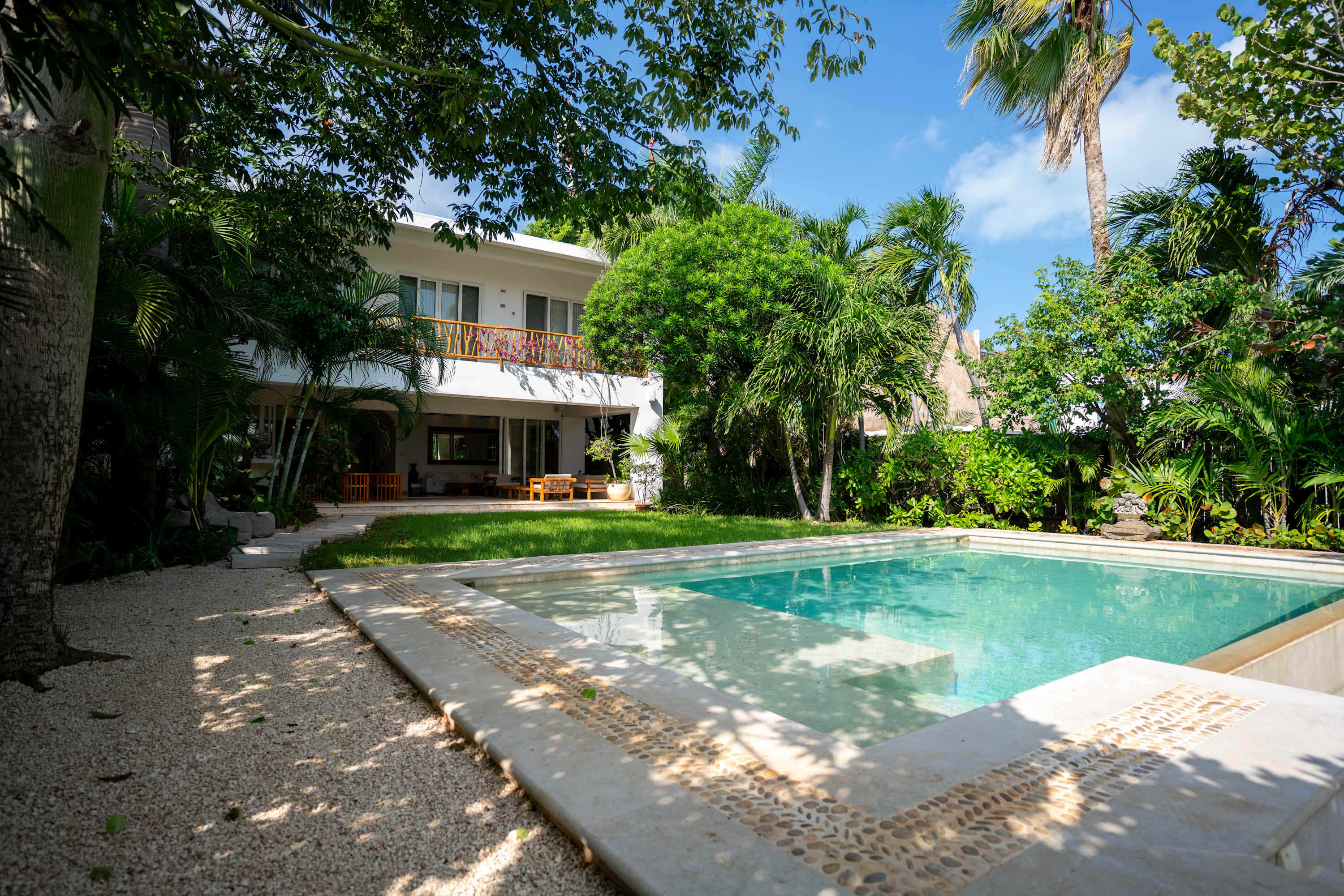 Modern white house with balcony, surrounded by lush greenery and a turquoise swimming pool.