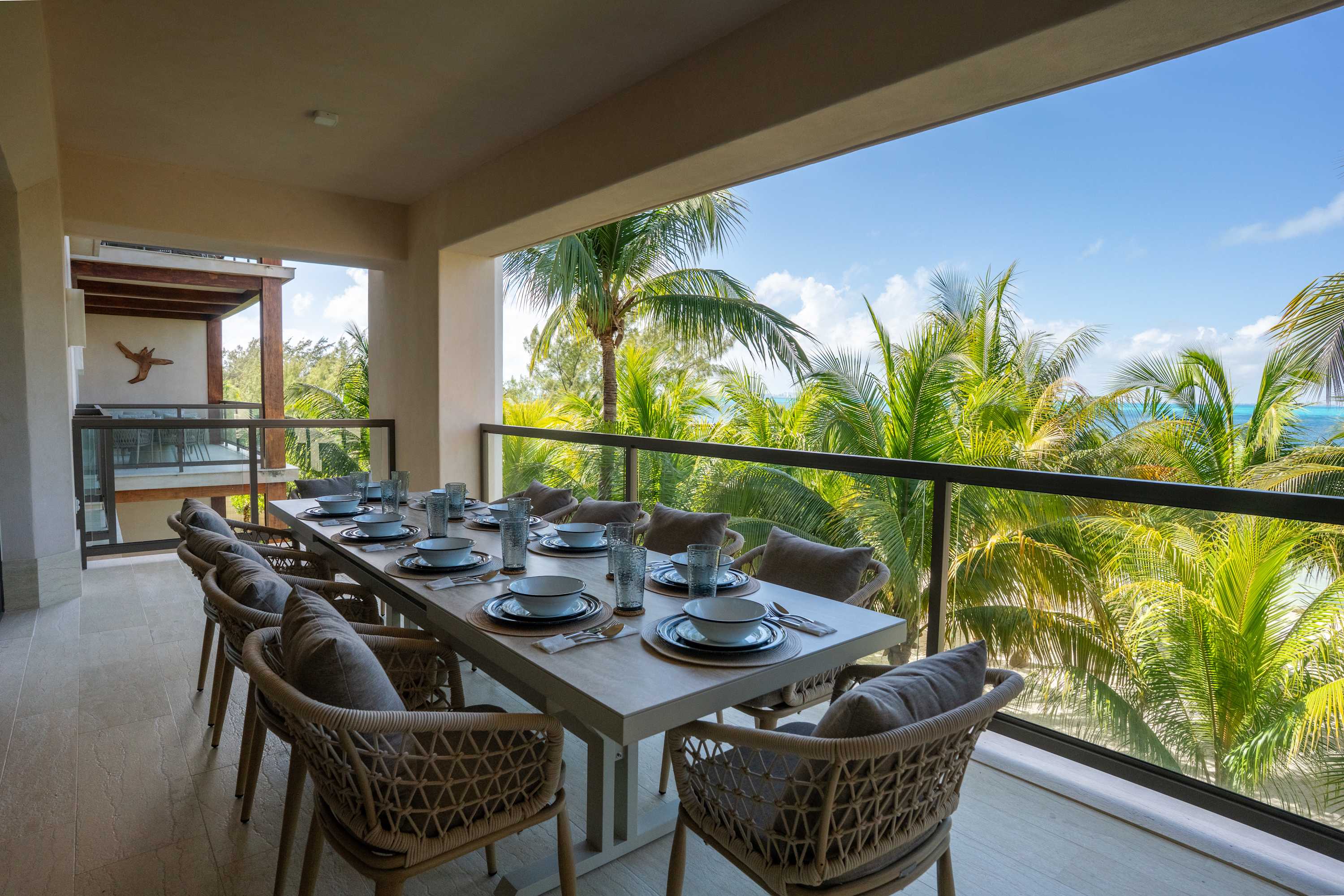 A covered balcony dining area with a table set for ten overlooking palm trees and the ocean.