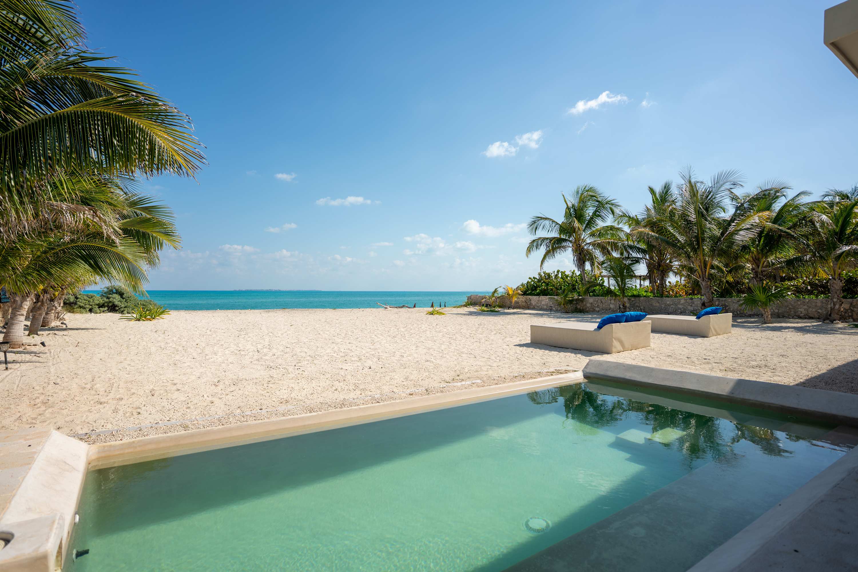 A tranquil pool and sandy beach with palm trees under a clear blue sky.