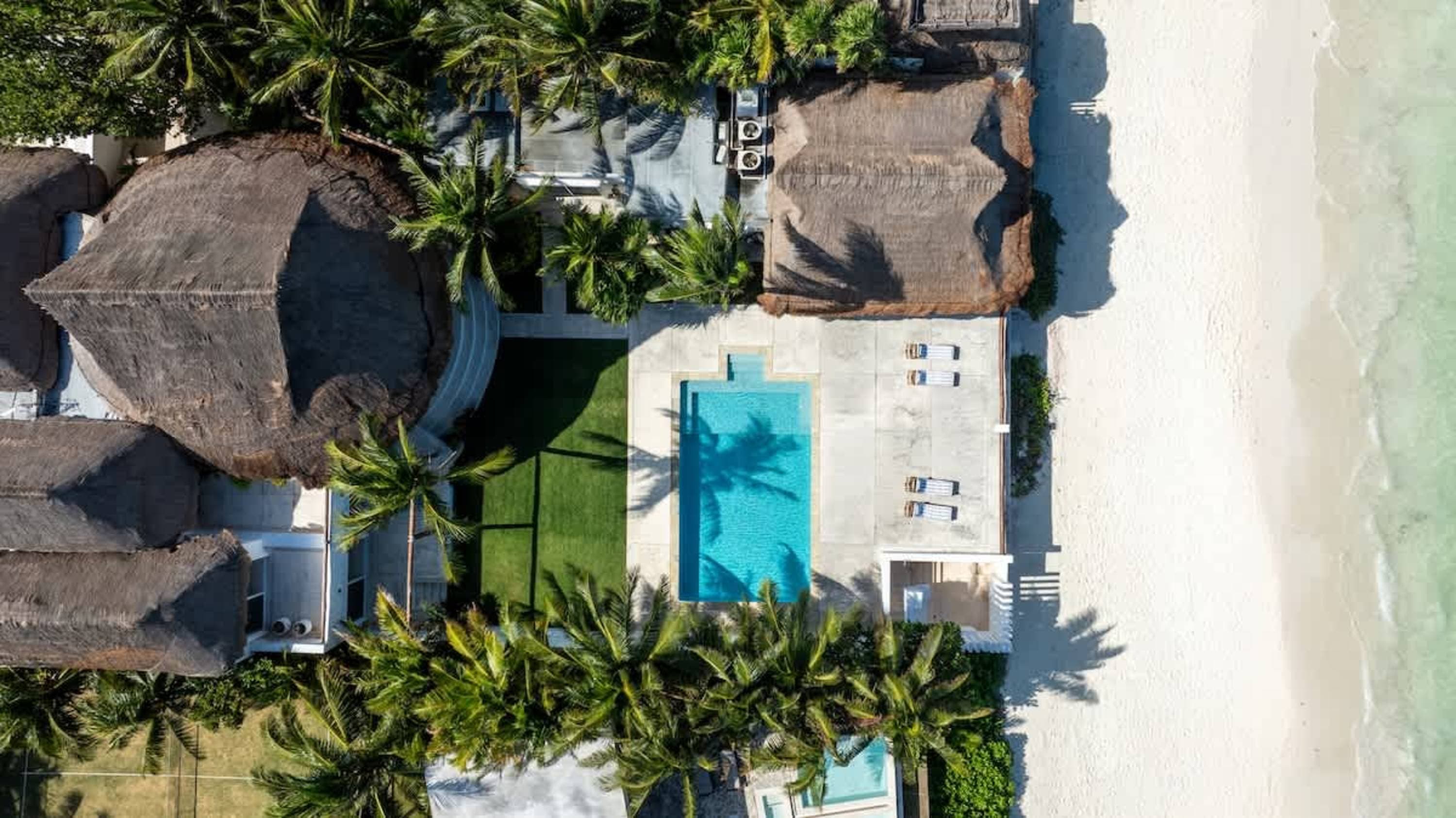 Aerial view of a beach resort with thatched roofs, a swimming pool, and palm trees.