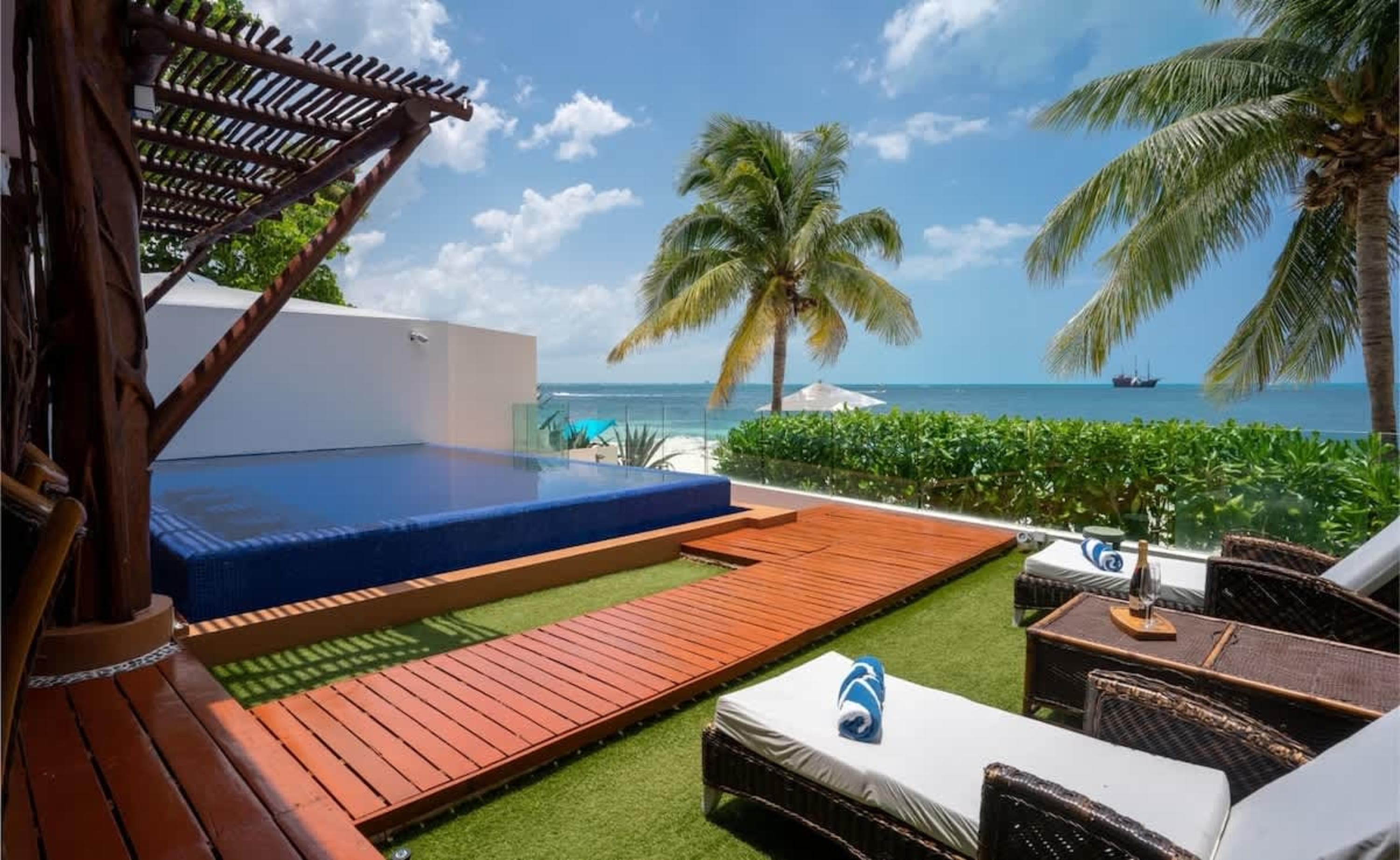 A luxurious beachfront patio with a pool, lounge chairs, and ocean views under a bright blue sky.