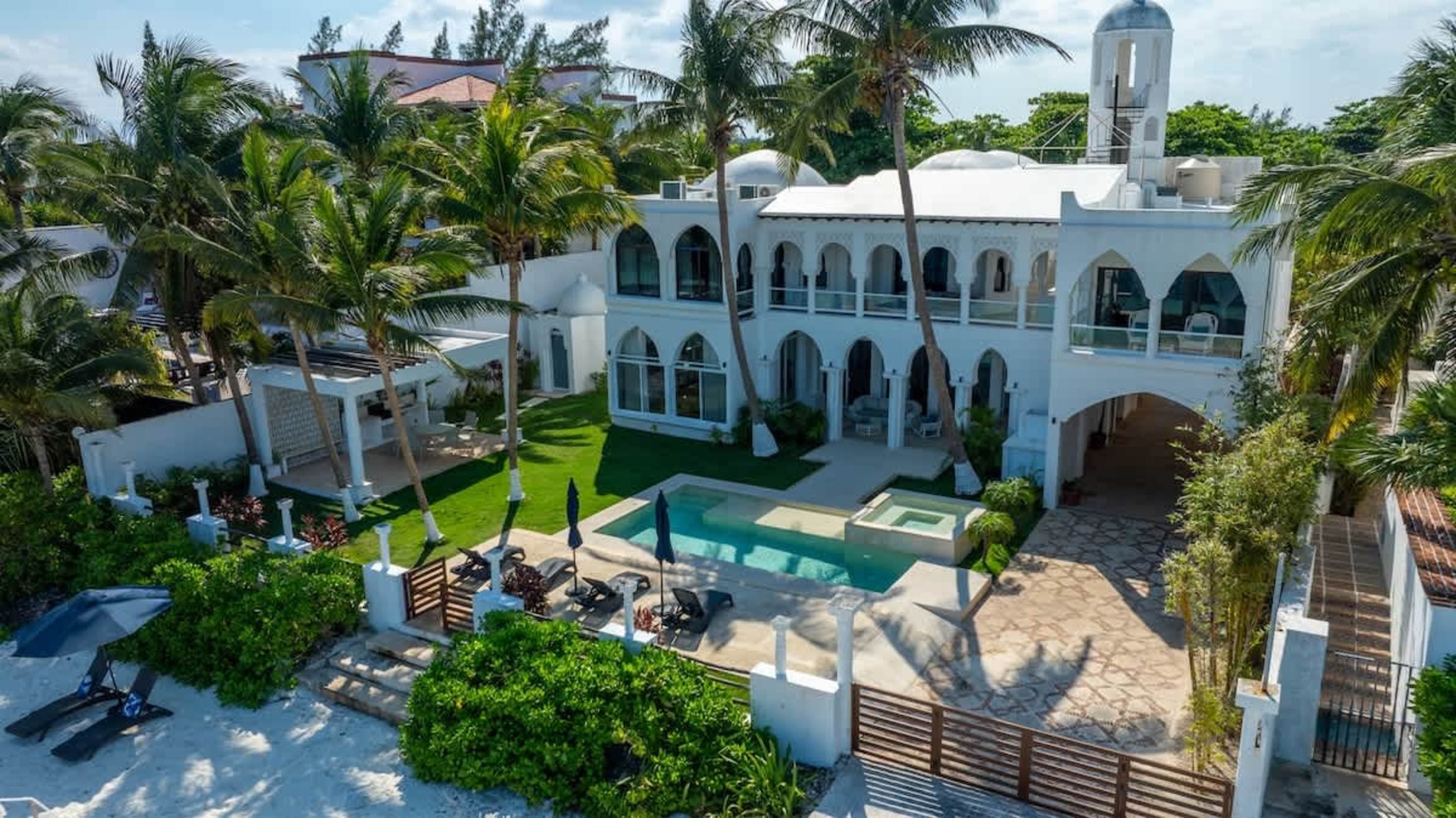 A white villa with a pool, surrounded by palm trees, and a sandy beach.