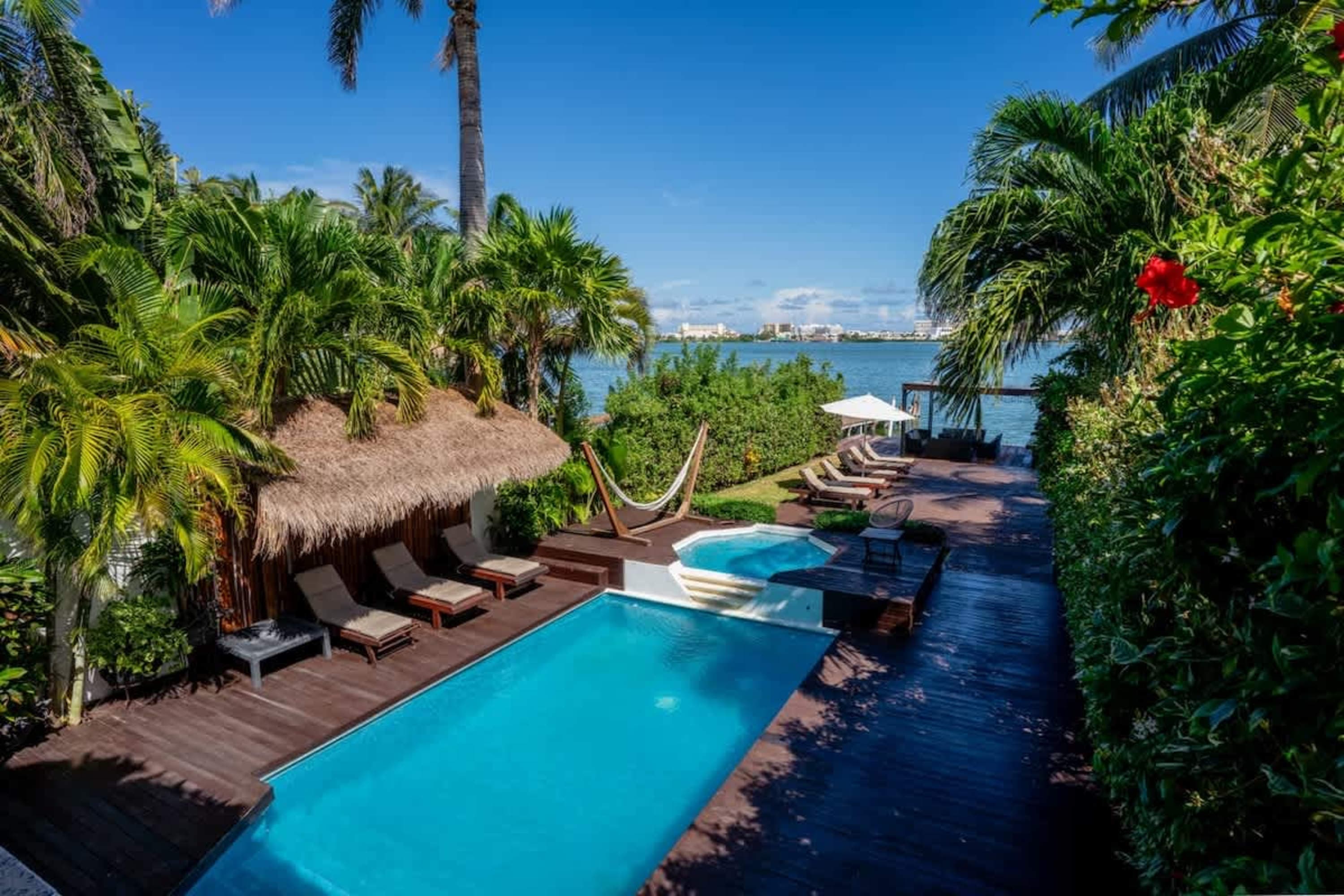 A tropical poolside with loungers, a thatched cabana, and a hammock overlooks a calm bay.